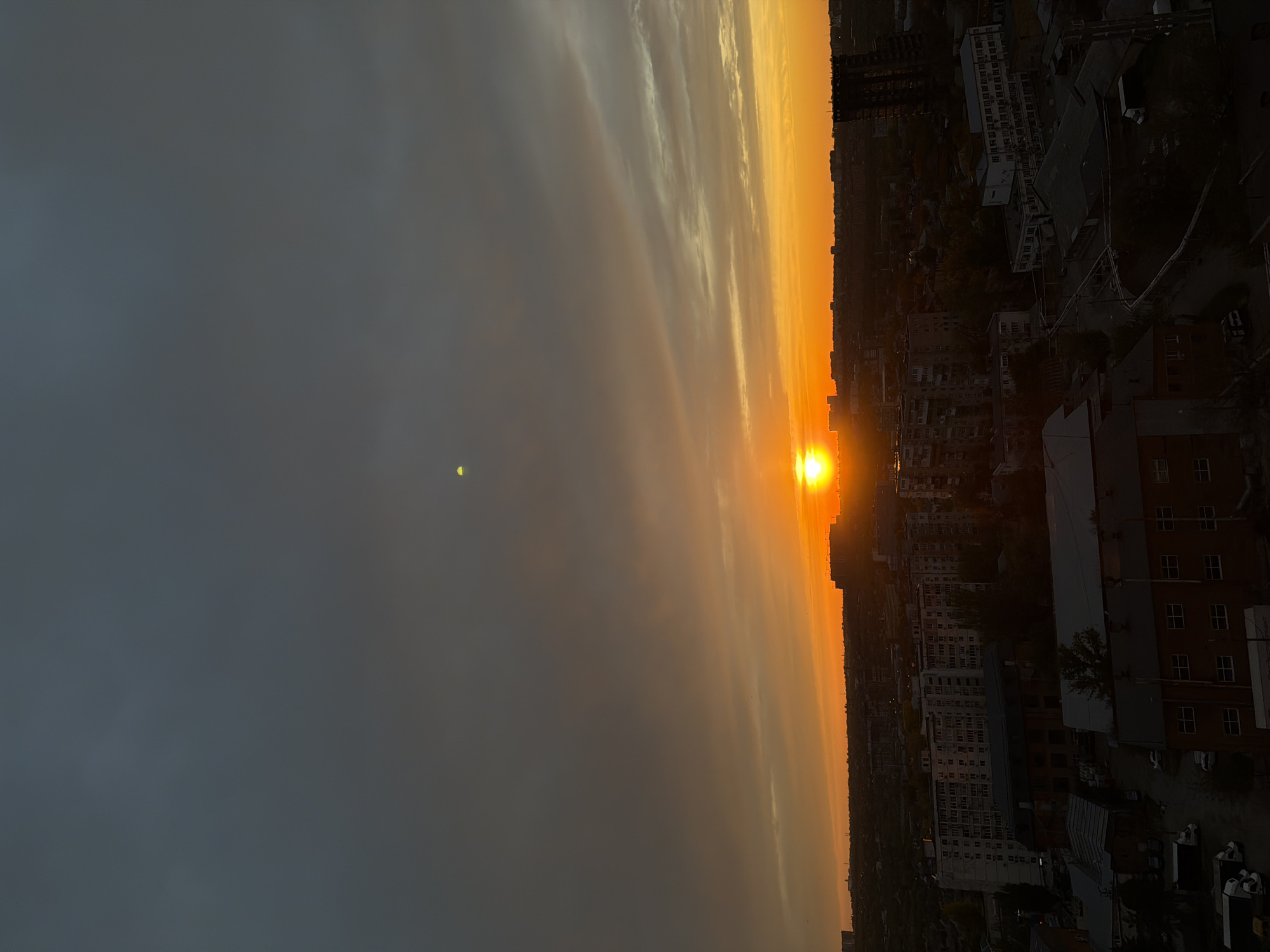 The image shows a sunset over the city of Rostov-on-Don, Russia. The sun is positioned on the horizon, casting a bright orange glow across the sky and illuminating the buildings below. The sky is a gradient of colors, transitioning from dark gray on the left to bright orange near the horizon. The buildings of the city are visible on the right side of the image, appearing as dark silhouettes against the bright sky. No people are visible.
