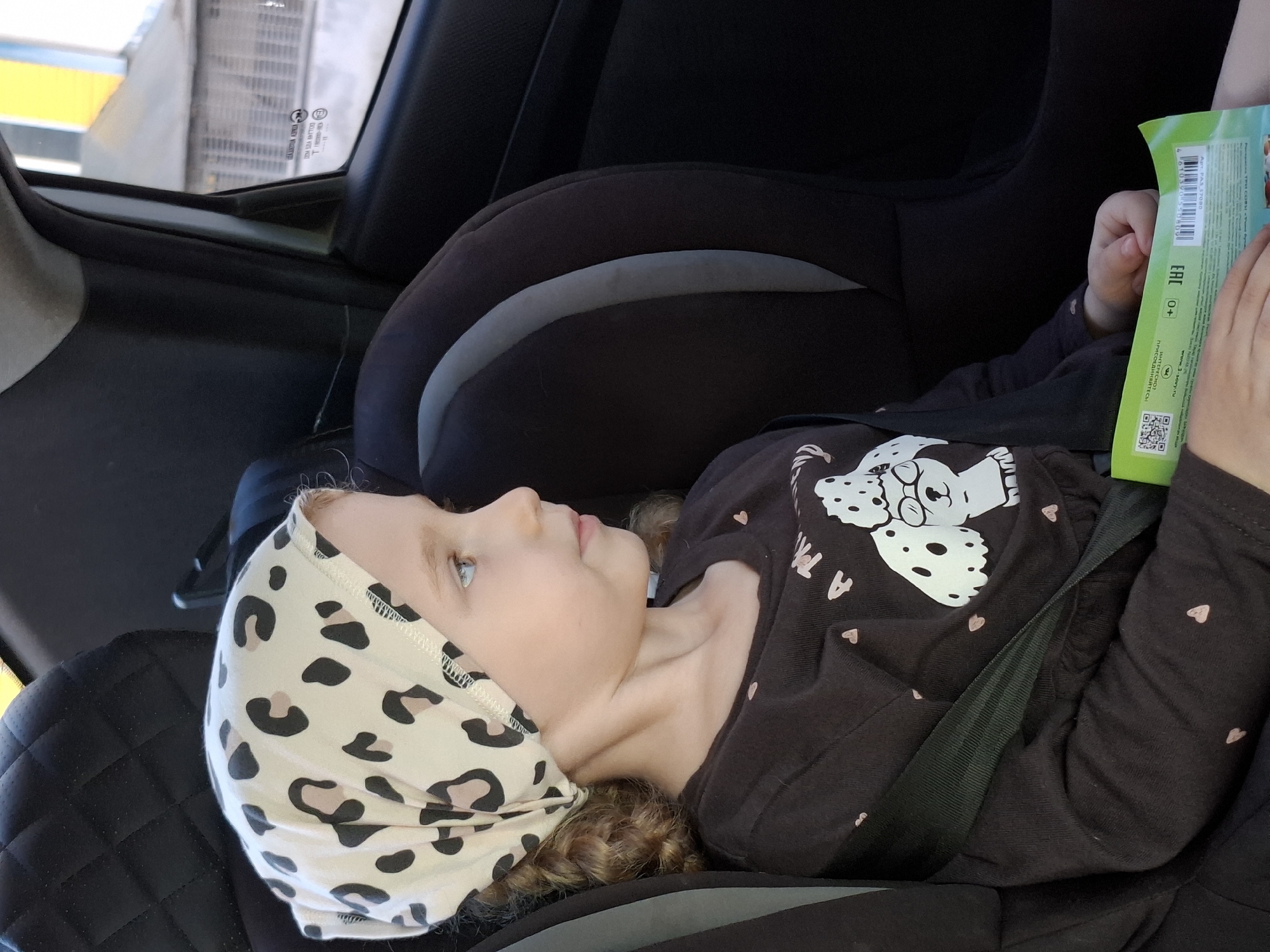 A young girl is seated in a car seat. She wears a patterned head covering and a black long-sleeved shirt with a Dalmatian design. The girl is buckled in with a seat belt and is holding a green package with text in a Cyrillic alphabet. The location is the city of Chaikovsky, Russia, as indicated by the Cyrillic text on the package.