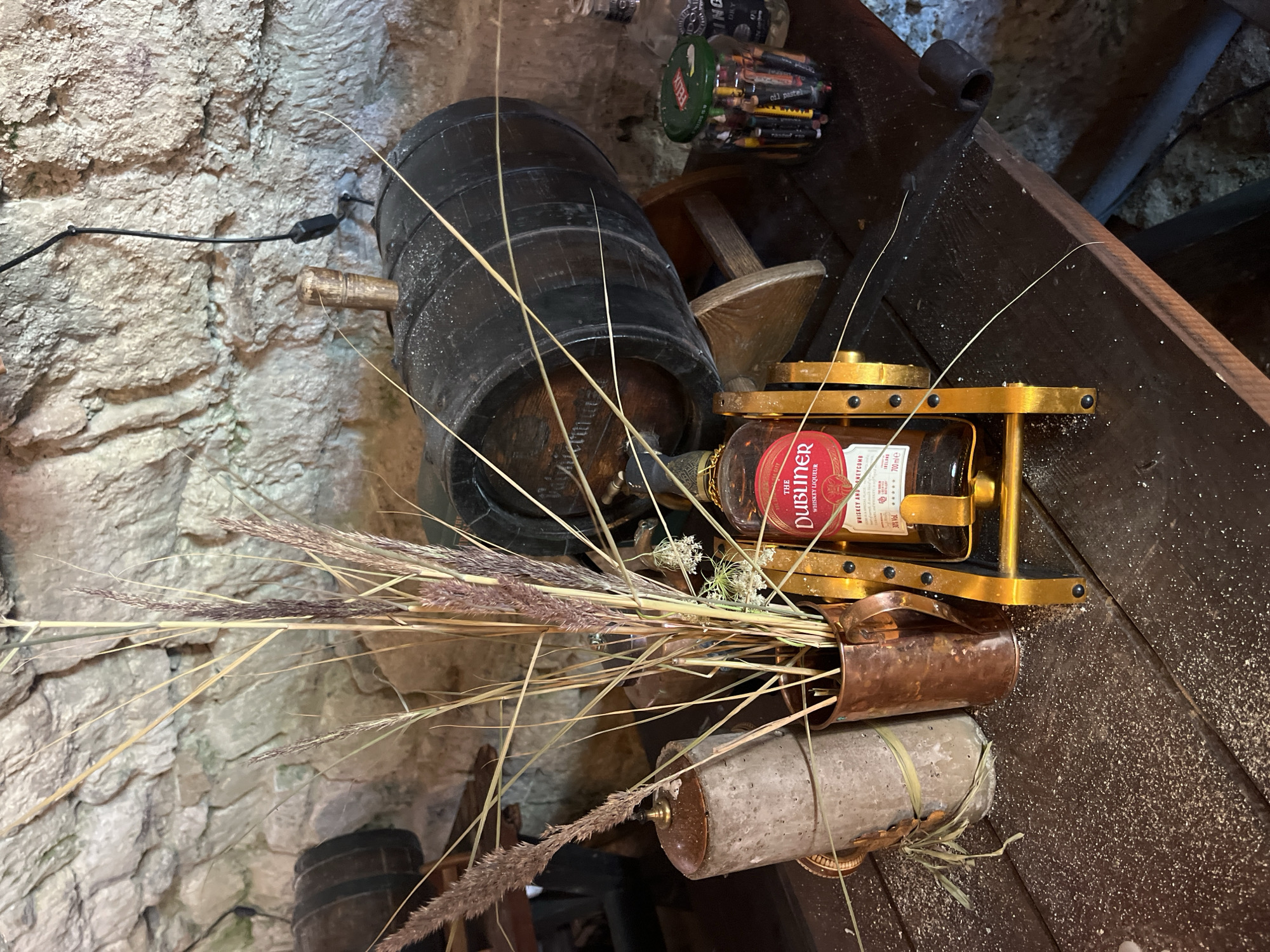 The image shows a close-up view of a rustic table setting. In the foreground, there is a bottle of "The Dubliner" whiskey liqueur displayed on a golden stand. Next to it, a copper mug holds a bouquet of dried grasses and a delicate white flower. To the left, a tall, cylindrical object, possibly a candle or a decorative piece, is wrapped with what appears to be straw. The table surface is dark wood, covered in a fine white powder, possibly sawdust or salt. In the background, a dark wooden barrel rests on a stand, against a rough stone wall. A dark electrical wire snakes down from the wall to the barrel. A glass jar filled with colored pencils, its lid featuring the brand "Felix", is visible to the right of the barrel. The overall impression is one of an aged, possibly cellar-like or traditional pub atmosphere. The presence of the barrel and stone wall suggests a historical setting, and while no specific landmarks are visible, such an environment might be found in older establishments within Kuressaare, Estonia, known for its historical architecture and castle.
