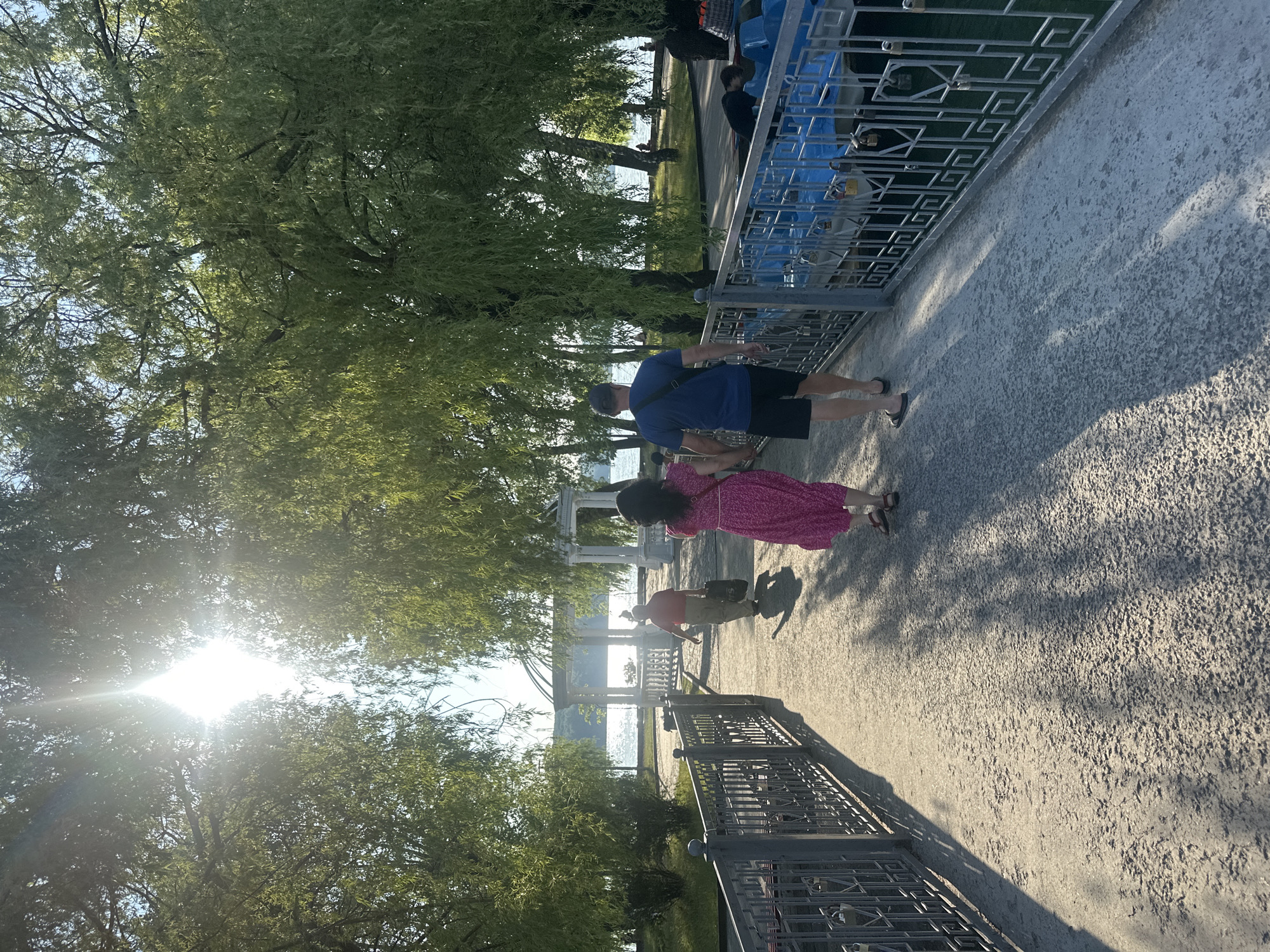 A sunny day scene at the Ternopil Lake embankment in Ternopil, Ukraine. A paved pathway, bordered by a decorative metal fence, leads away from the viewer. Two people are walking on the path, a man wearing a blue t-shirt and black shorts, and a woman in a pink dress. In the background, a woman in a red top and light pants walks further ahead, carrying a bag. Lush green trees with drooping branches line the pathway. Across the pathway, a body of water is visible, with a gazebo structure on the embankment and several blue paddle boats docked along the edge. Sunlight streams through the trees, casting dappled shadows on the path.