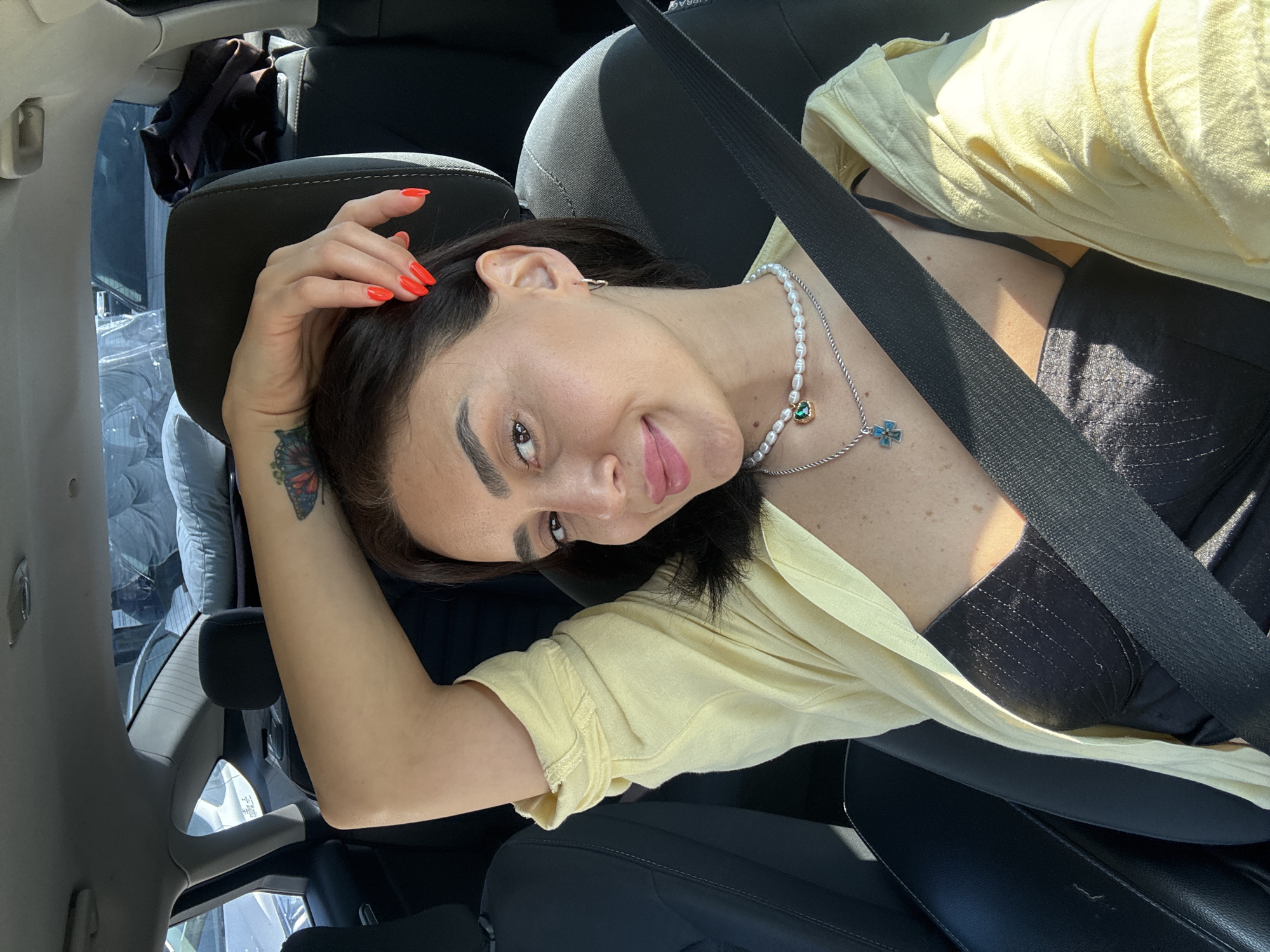 A woman with dark hair, wearing a black bustier, a light yellow open shirt, and a pearl necklace with a green pendant, is seated in the passenger seat of a car. She is smiling and has her arm raised, resting her hand on the back of her head. The seatbelt is fastened across her chest. The interior of the car is visible, with black seats and a glimpse of the car's headliner. Sunlight streams in through the windows, casting shadows on the woman and the car's interior. The background shows a blurred view of what appears to be a parking lot with other vehicles and possibly some outdoor structures. No specific details of Tbilisi, Georgia, are discernible from the image.