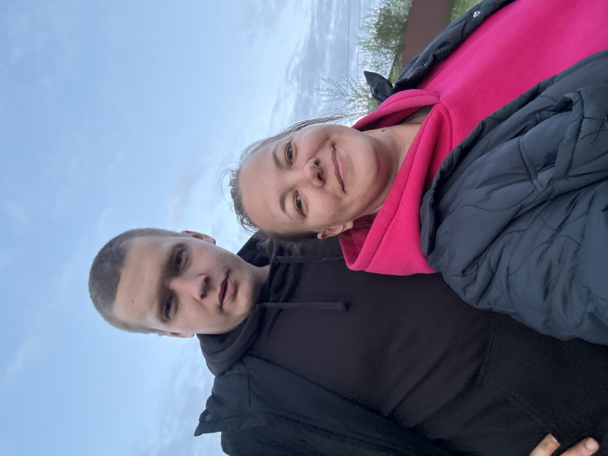 Two individuals, one male and one female, are positioned close to each other in an outdoor setting under an overcast sky. The woman, likely in her 30s or 40s, is wearing a pink hooded sweatshirt and a dark jacket. The man, potentially in his 20s or 30s, is wearing a black hooded sweatshirt and a dark jacket. The image suggests a selfie taken at an angle, looking upwards towards the sky. Vegetation, possibly trees, is visible in the background. The image was taken outside, possibly in Dmitrov, Russia. The exact location and the specific actions or interactions between the individuals are not discernible from the image.