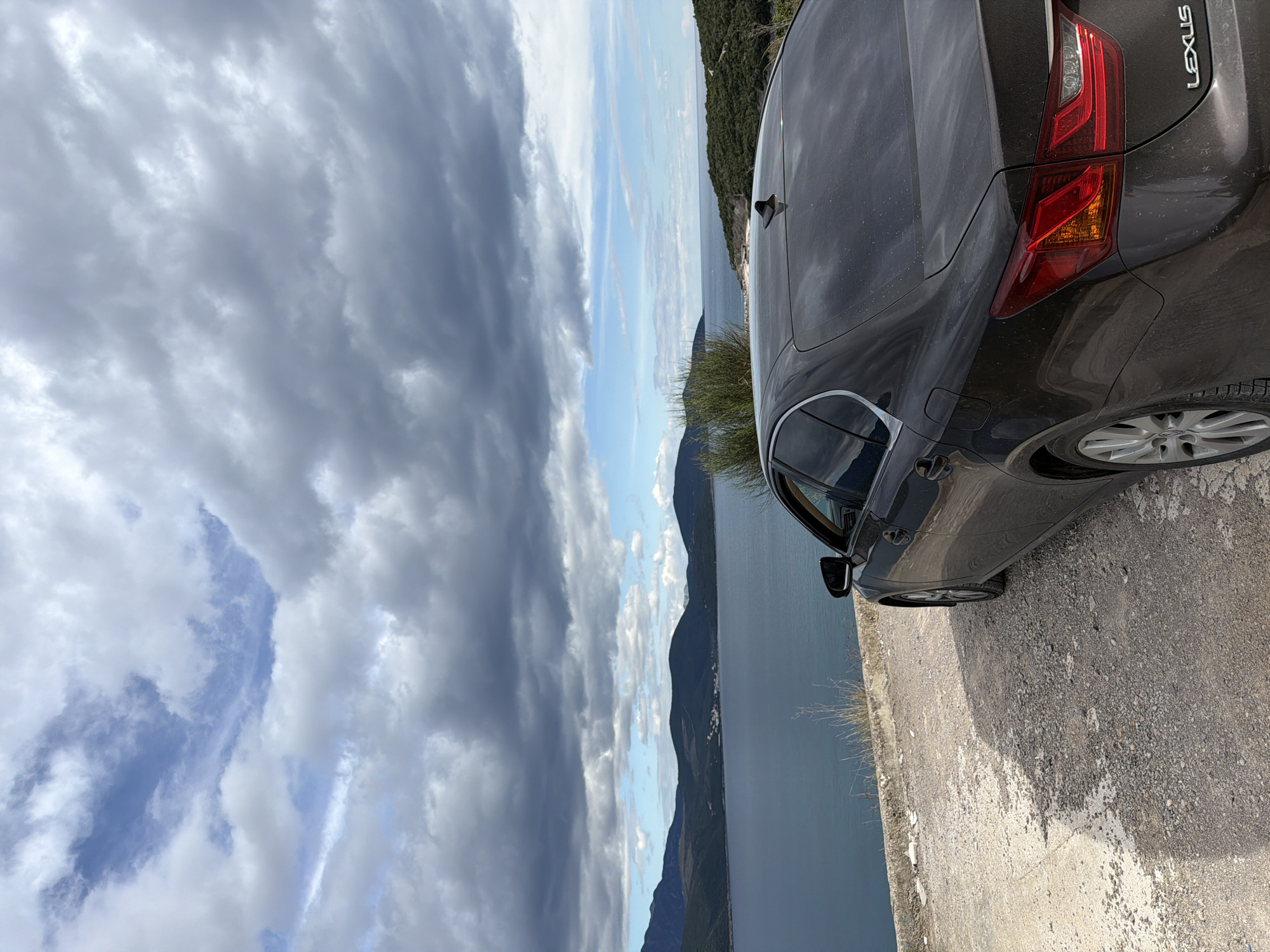 The image shows a gray Lexus sedan parked on a concrete road near the water's edge. The sedan occupies the right portion of the scene, with its rear facing the viewer. The rear light, tire, and part of the car body are visible. To the left is a view of the bay with mountains in the distance and a cloudy sky above. The photo was likely taken near the city of Radovići, Montenegro.