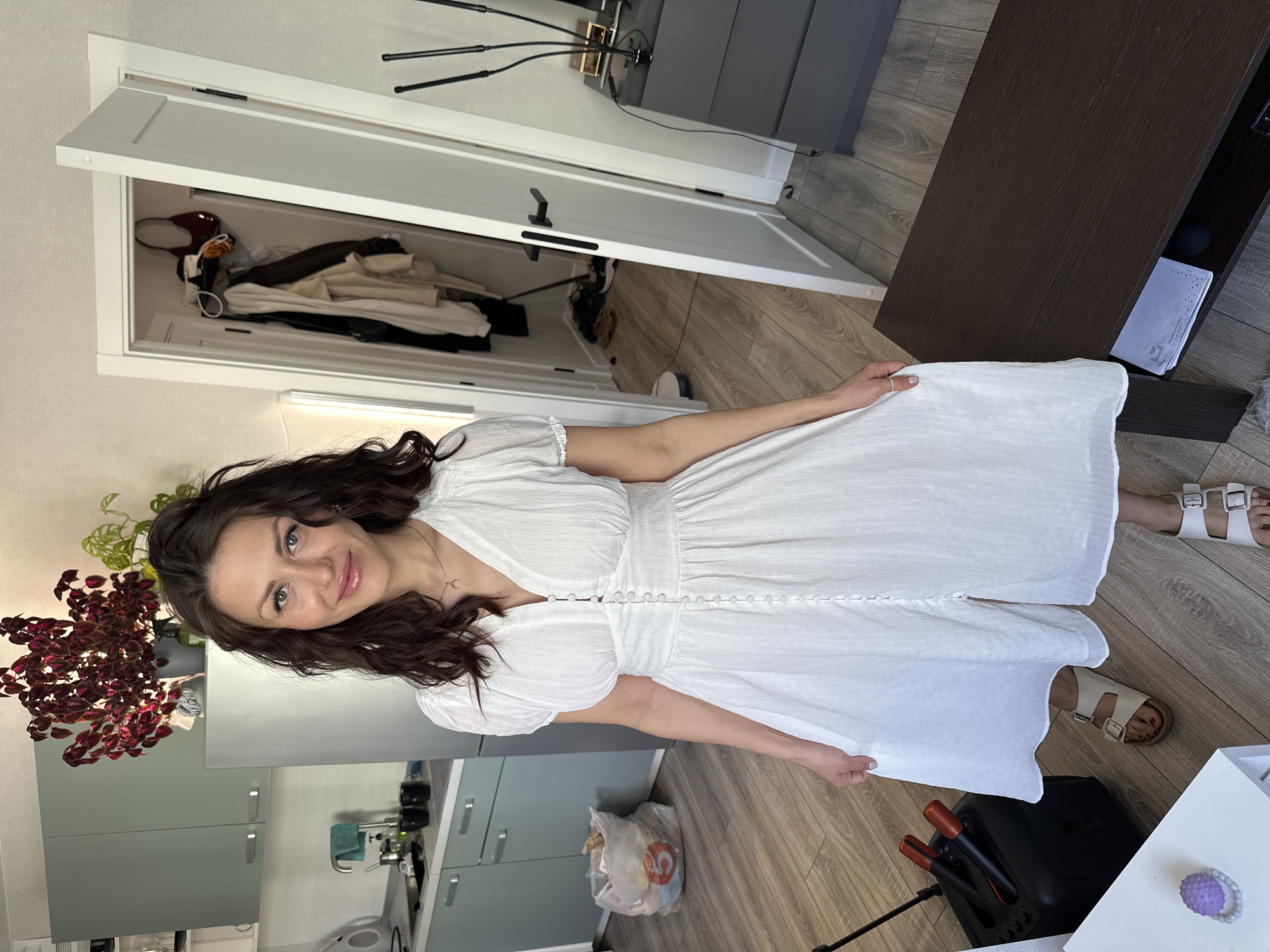 A woman in a white dress stands in a room. The room has wooden floors, light green kitchen cabinets, and a brown TV stand. A closet with white doors is partially open, revealing clothes inside. A potted plant with red leaves sits on a counter. A black karaoke machine and a shopping bag are on the floor. The location is Murino, Russia.