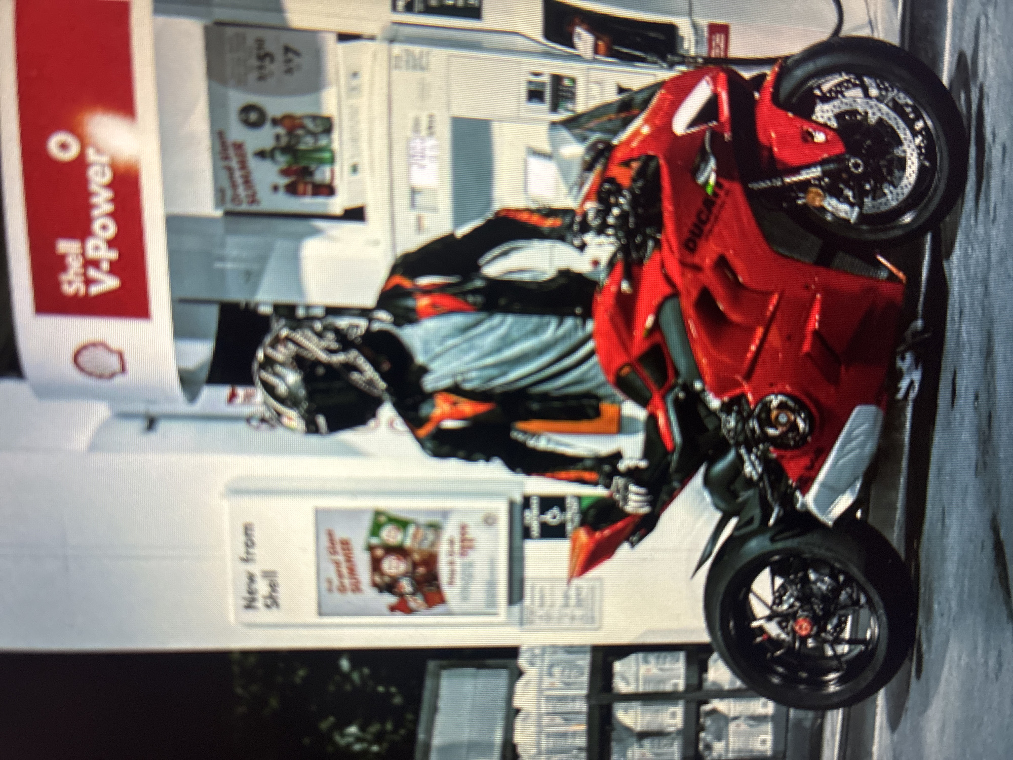 The image captures a red Ducati motorcycle and its rider at a Shell gas station in Asenovgrad, Bulgaria, during daytime. The motorcycle, likely a sportbike model, is prominently displayed in the foreground, featuring bright red fairings, black wheels with intricate brake discs, and partial "DUCA" branding on its side.
Standing next to the motorcycle is a person dressed in full protective motorcycle gear, including a black helmet, a black, orange, and grey jacket, and gloves. The activity appears to be a stop at the gas station, possibly for refueling or a brief break.
In the background, multiple gas pumps are visible, adorned with the iconic red and yellow Shell branding. A large "Shell V-Power" sign is prominent on the gas station canopy structure. Advertising posters on the pumps display text such as "New from Shell," "Grand Slam SUMMER," and other "SUMMER" promotions. The numbers "5" and "7" are also visible, possibly indicating prices or special offers. The scene is brightly lit, suggesting clear, sunny weather during the day.