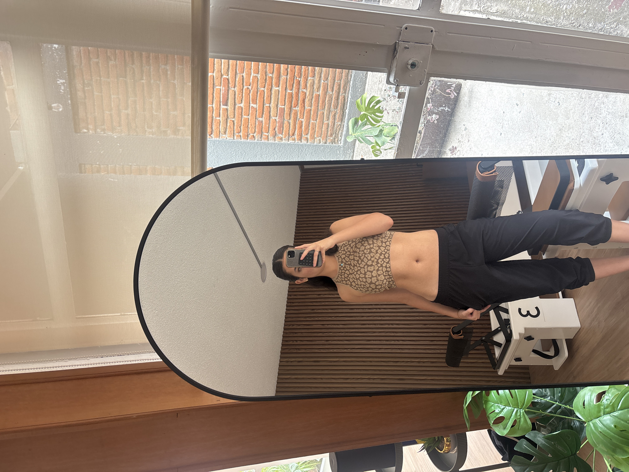 A woman is taking a mirror selfie in a room in Puebla, Mexico. The room has a wooden floor and blinds on the windows. A brick wall is visible through the window, as is a plant outside. Inside the room, there's a pilates reformer and a potted plant. The woman is wearing a leopard print top and black pants.