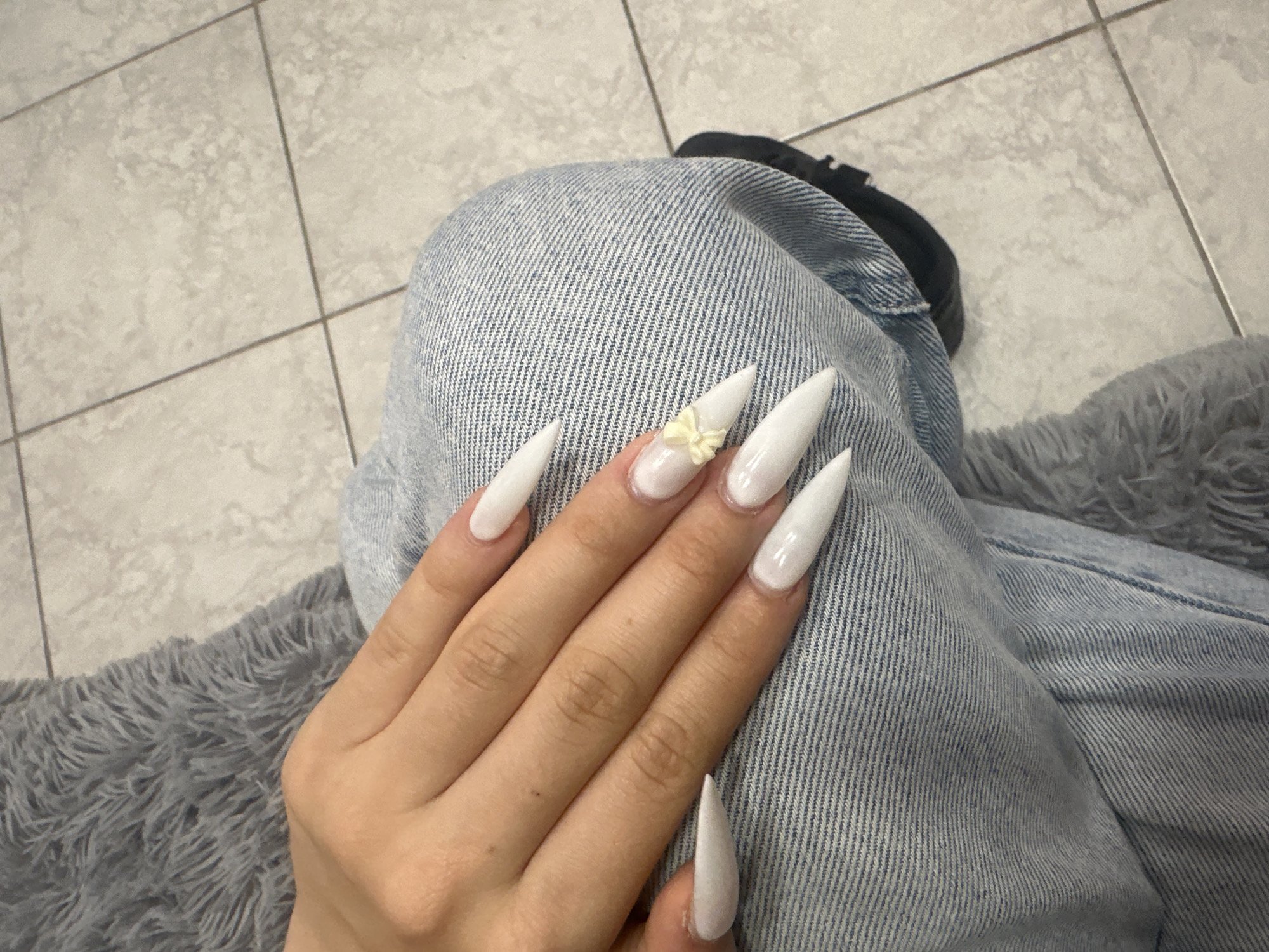 The image shows a close-up of a person's left hand, which has long, pointed, white acrylic nails. One of the nails, on the ring finger, has a small, light yellow, three-dimensional butterfly charm attached. The hand is resting on a person's lap, who is wearing light blue denim jeans. Behind the lap, a grey, fluffy blanket is visible. The background consists of light-colored tiled flooring. The image is cropped to focus on the hand and the immediate surroundings. This scene is not identifiable as being located in the city of Stamboliyski, Bulgaria.