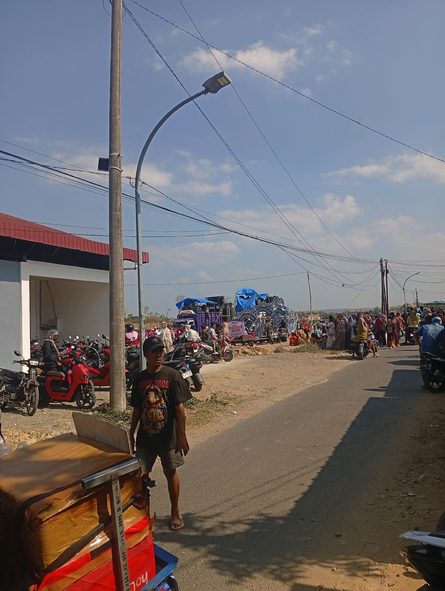A street scene in Nglojo, Indonesia, shows a gathering of people on a sunny day. A man in a black t-shirt with a Mario Bros. graphic and shorts stands in the foreground, facing right, near a cart with red and brown boxes. Behind him, a street light pole with electrical wires extends upwards. A building with a red roof and white walls is visible to the left, with several red and silver motorcycles parked in front of it. Further down the street, a crowd of people are gathered around what appears to be a stage or performance setup covered with blue tarps, with more motorcycles parked nearby. The road is paved, with dirt and vegetation on the sides. The sky is blue with scattered white clouds.