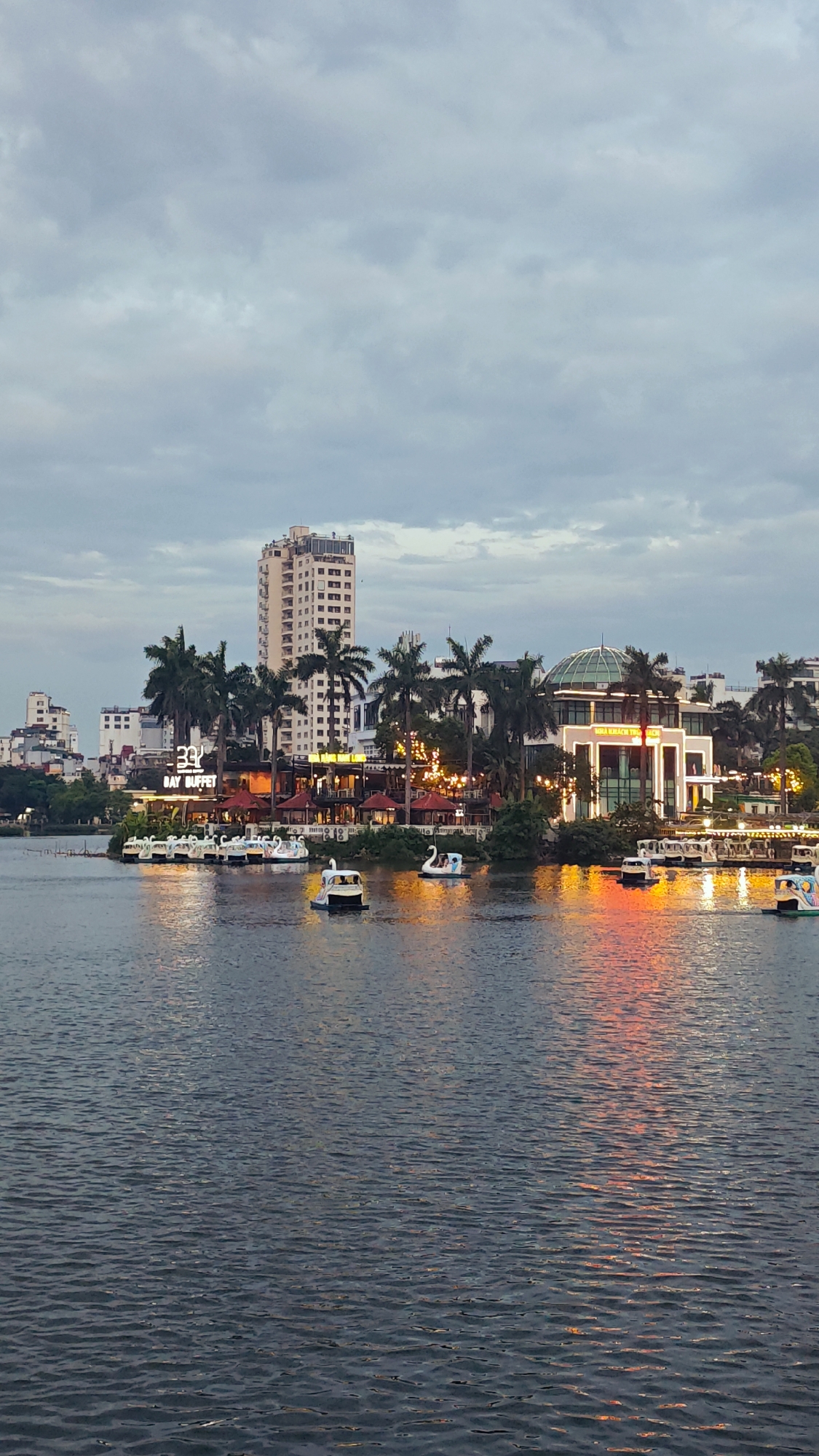 The image captures a waterfront scene at dusk in Hanoi, Vietnam, likely near West Lake, given the presence of swan-shaped paddle boats and illuminated buildings lining the shore. A body of water, reflecting the orange glow of lights from the buildings and the muted grey of the overcast sky, dominates the foreground. Several white swan-shaped paddle boats are scattered across the water, with one in the foreground being paddled. Behind the water, a row of buildings is visible, some featuring brightly lit signage and architectural lighting, including a prominent building with a domed glass roof. Palm trees are interspersed among the buildings, suggesting a tropical or subtropical climate. A tall, slender skyscraper stands further in the background, piercing the cloudy sky. The overall atmosphere is serene, with the transition from day to night marked by the artificial lights beginning to illuminate the cityscape.