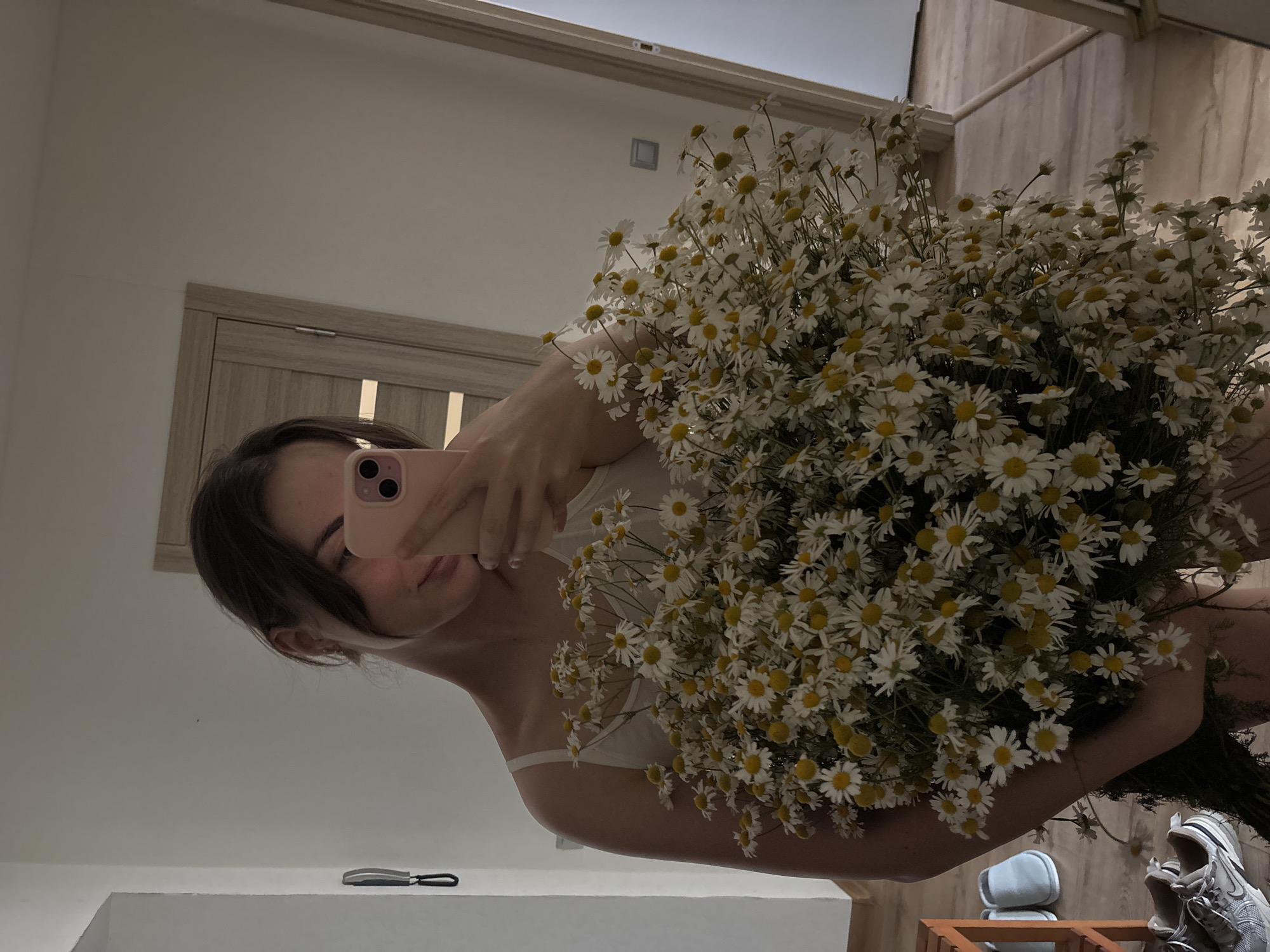 A young woman is taking a mirror selfie in a room. She is holding a large bouquet of daisies in her arms and is looking down at her phone, which is held up to take the picture. The woman is wearing a white tank top. The room appears to be a bedroom or dressing area, with a door and part of a closet visible in the background. A wooden shelf with a pair of blue slippers and a pair of sneakers is also visible in the foreground. The lighting in the room is soft and natural.
