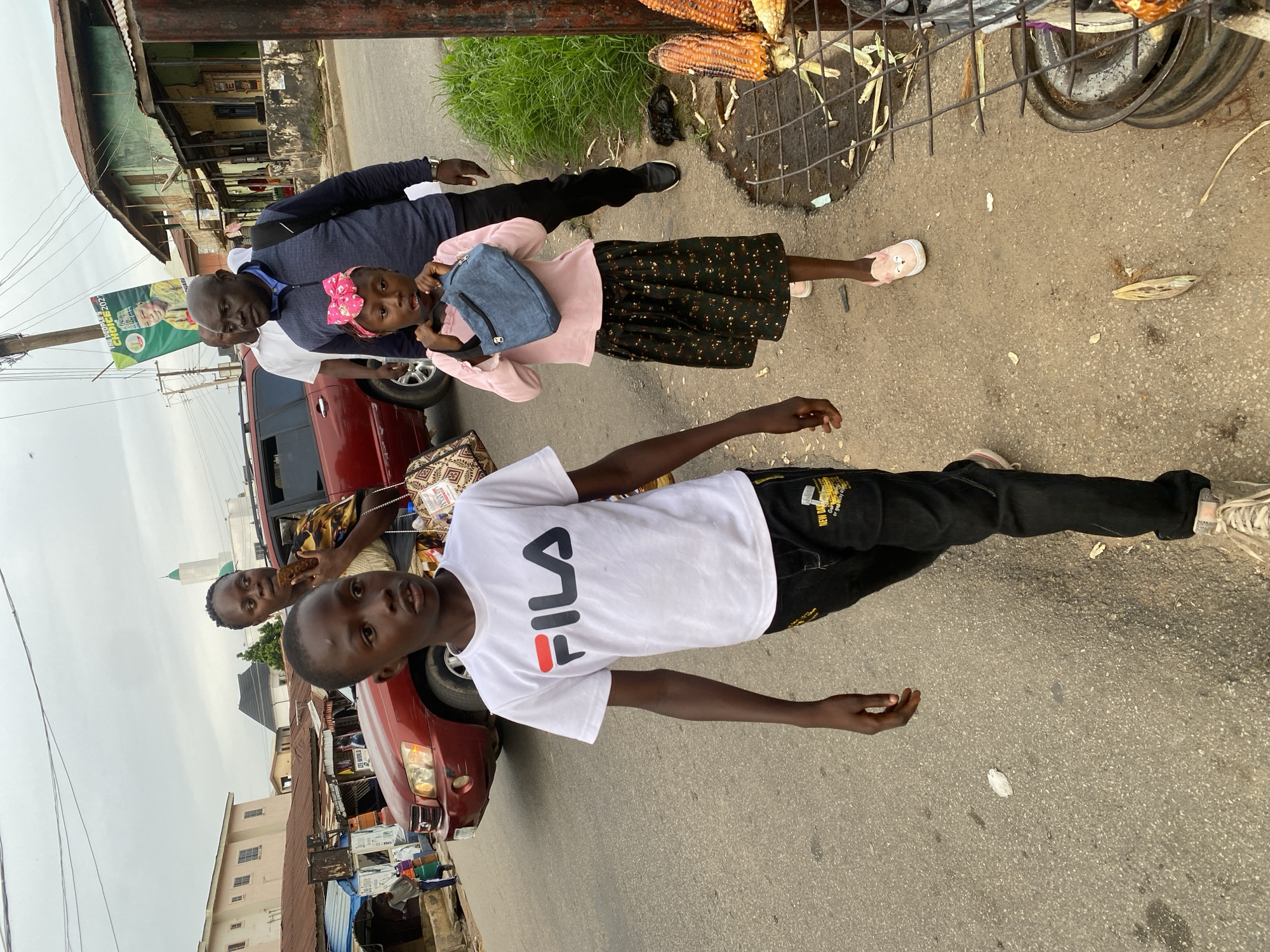 The image depicts an outdoor scene in Jos, Nigeria, likely on a street. In the foreground, a young boy wearing a white Fila t-shirt and black pants stands, looking towards the camera. Behind him, a red car is parked. To the left of the car, an adult man in a blue sweater and a woman in a pink top and floral skirt are partially visible. The woman is holding a young girl wearing a pink bow in her hair and a denim backpack. Another person's arm is visible reaching out from the right side of the frame. In the background, there are buildings, some appearing weathered, and a green banner with text that is difficult to read clearly. There are also some corn cobs on a metal grate, suggesting a market or roadside stall. The lighting suggests it might be daytime, possibly overcast. The overall impression is a candid moment of people gathered on a street, with the presence of a car and roadside vendors hinting at a busy, everyday environment.