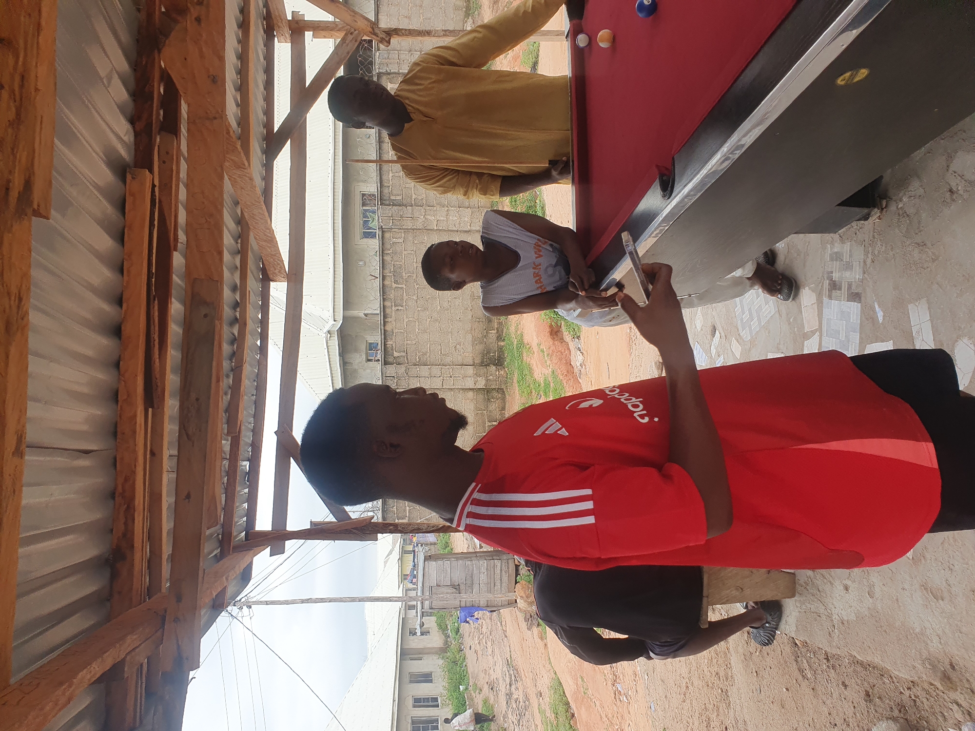 In Lokoja, Nigeria, under a rustic, open-air wooden shelter with a corrugated metal roof, a group of people are gathered around a red pool table. A young man in a red t-shirt stands to the left, holding a mobile phone. To his right, a person in a white tank top leans on the pool table, looking at the game. Behind them, another man in a mustard-yellow tunic holds a pool cue, appearing to be playing. The ground is a mix of sand and concrete tiles, with some distant buildings and a wire fence visible in the background. The atmosphere seems casual, with a sense of community and recreation.