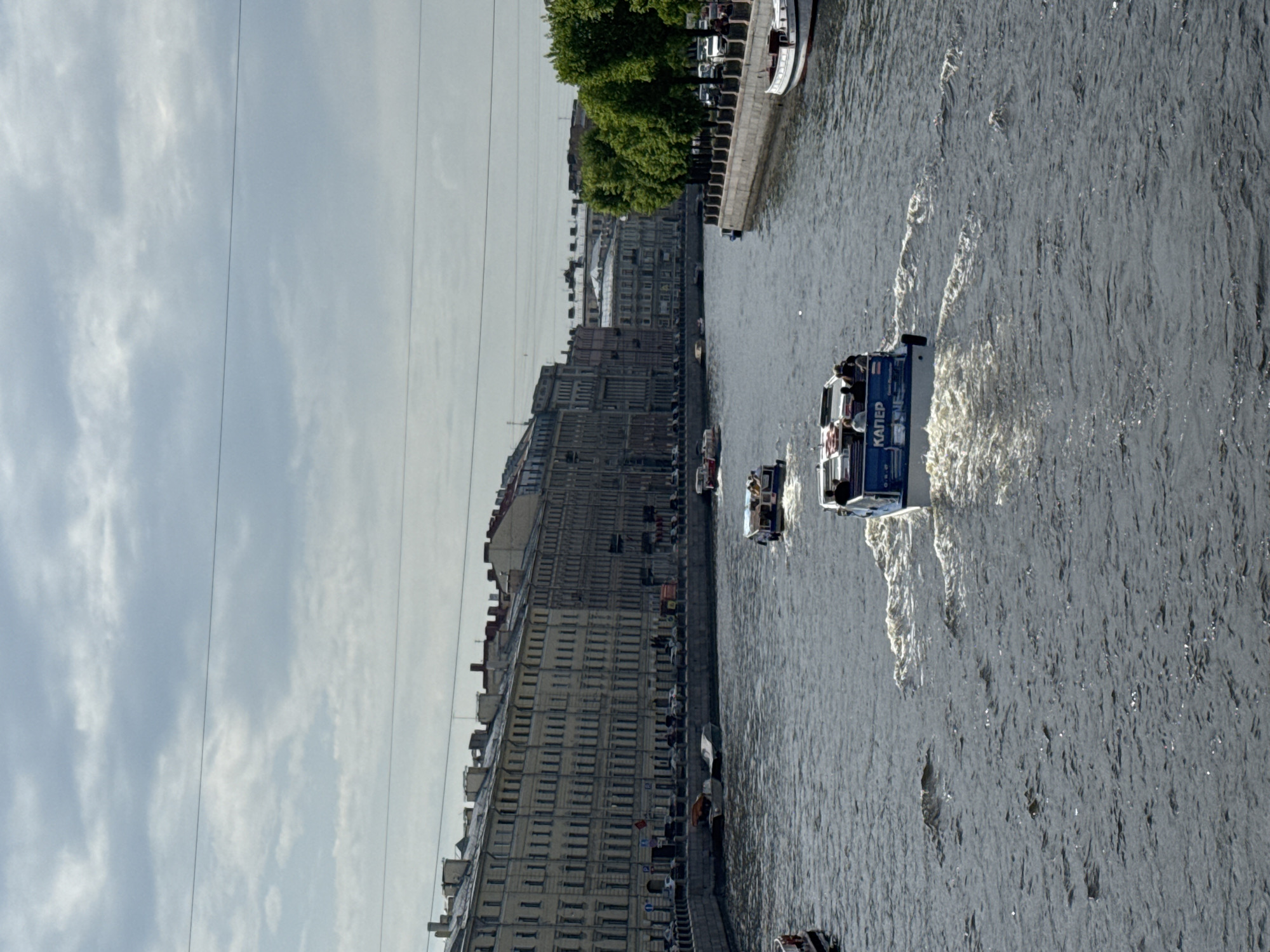 A view of the Fontanka River in Saint Petersburg, Russia, features several river boats navigating the water. Along the riverbank, the historic architecture of the city lines the quay, with the facades of buildings visible. Onboard one of the tour boats, identifiable human figures indicate it is carrying passengers. The river reflects the sky and the buildings, while the movement of the boats causes ripples and wakes on the water's surface. Overhead, utility lines run parallel to the river, and trees can be seen on the other bank.