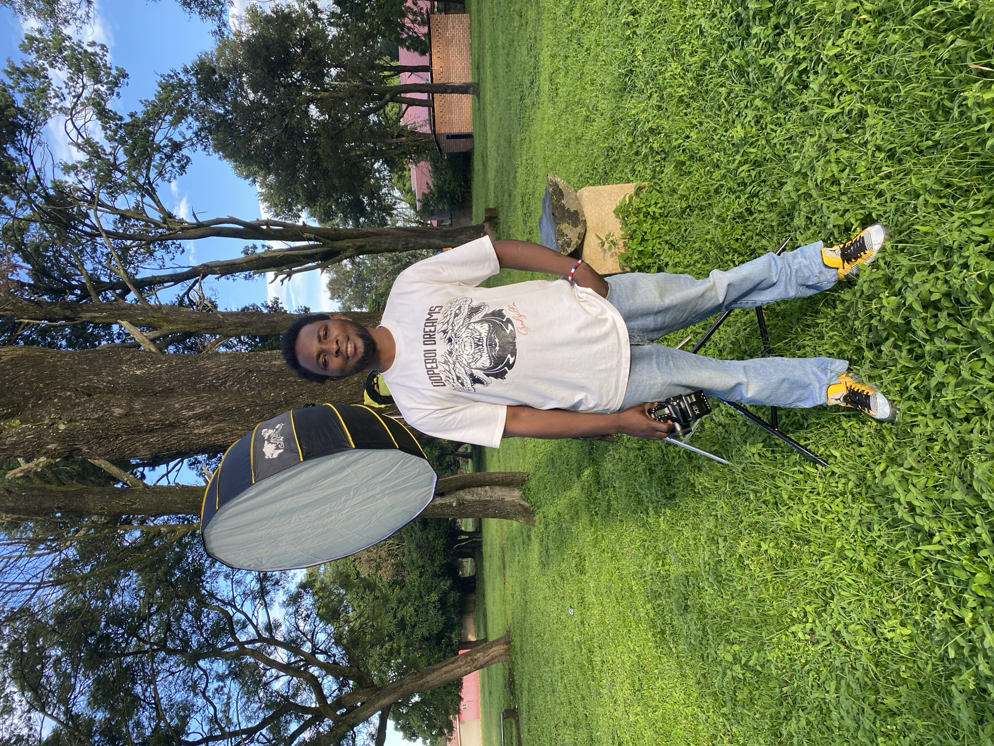 A young Black male is standing in a grassy outdoor area, possibly a park or school grounds. He is wearing a white t-shirt with a graphic design and blue jeans, paired with yellow and black sneakers. He is holding a camera in his right hand, with his left hand in his pocket. To his left, a large photography lighting umbrella is set up on a tripod. In the background, several trees are visible, along with a building with a red roof and pink walls. The sky is blue with scattered clouds. The scene appears to be an outdoor photography setup in an urban or semi-urban environment, potentially within the city of Jos, Nigeria, given the architectural style and vegetation.