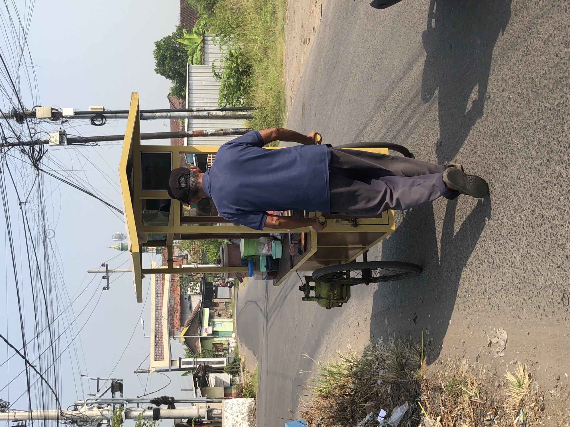 An adult male wearing a blue short-sleeved shirt, dark pants, a dark baseball cap, and brown shoes is pushing a yellow food cart on a paved road. The cart has a green propane tank attached to its side, two spoked wheels, and a canopy. The cart is laden with various containers and what appear to be food items. The man is walking away from the camera. The scene is set in Kesambi, Indonesia, with residential buildings, trees, and electrical poles visible in the background. The sky is clear and blue, indicating daytime. The road shows some tire tracks and debris along the side.