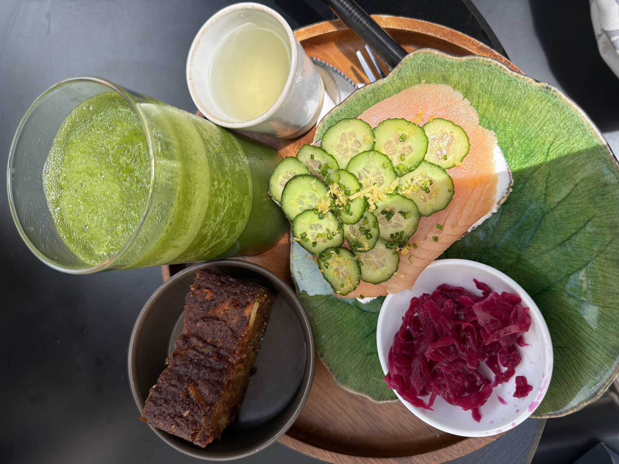 A top-down shot shows a wooden tray with a breakfast spread, likely in Tel Aviv, Israel, given the type of food presented. From left to right on the tray is a glass of green juice, a small bowl with a dark brown baked item (possibly a bread or cake), a cup of light yellow liquid, and a leaf-shaped plate containing a slice of salmon topped with sliced cucumbers and a sprinkling of green herbs, and a small white bowl filled with what looks like pickled red cabbage. A fork and knife rest on the tray near the leaf-shaped plate. The overall setting suggests a bright, sunny day.