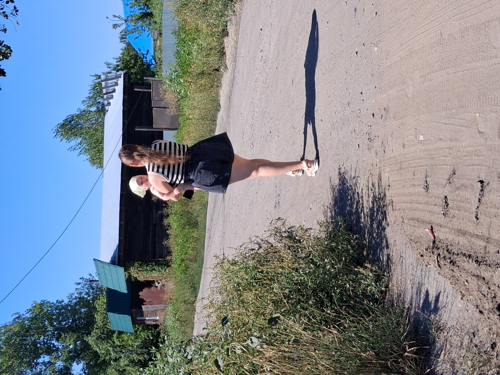 A woman, viewed from behind, carries a baby down a dirt road in the city of Semey, Kazakhstan. The woman has long, dark hair in a ponytail and wears a striped tank top and dark denim shorts. The baby is held against her chest and wears a light-colored head covering. They are walking away from the viewer towards a rustic, wooden building with a green, corrugated metal roof. The building is situated amongst overgrown vegetation and trees. The sky is clear and blue. The road is unpaved and shows tire tracks. Green brush grows on the left side of the road, and more vegetation is visible in the background. A shadow of the woman and baby is cast on the road to their right.