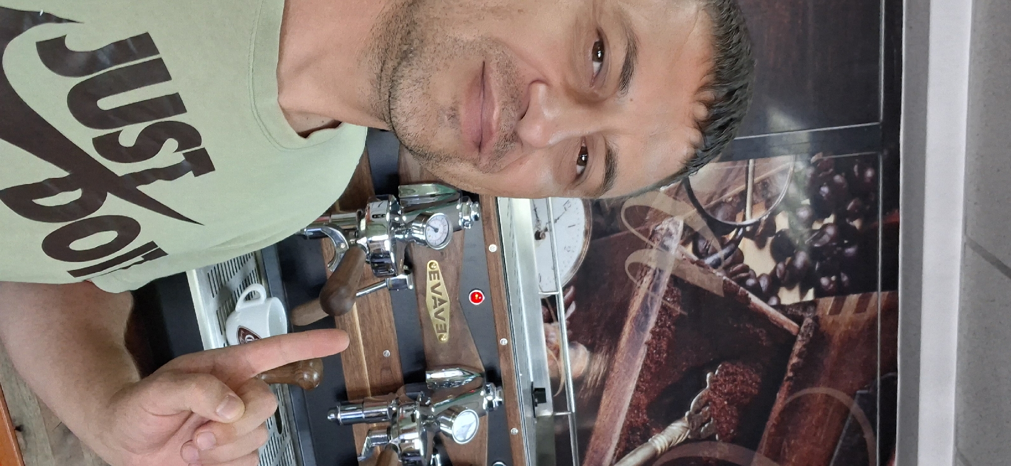A man is taking a selfie in front of an espresso machine. He is wearing a light green t-shirt with the Nike logo and the words "JUST DO IT" printed on it in dark brown. He has short, dark hair and is clean-shaven. He is smiling and holding up his right index finger. The espresso machine is made of stainless steel and wood, with two portafilters, pressure gauges, and a wooden handle. Behind the man, there is a mural or wallpaper depicting coffee beans and what appears to be a coffee grinder and scale. A small white espresso cup is visible on the drip tray of the machine. The environment appears to be an indoor cafe or coffee shop. The specific location within Chișinău, Moldova cannot be determined from the provided image.