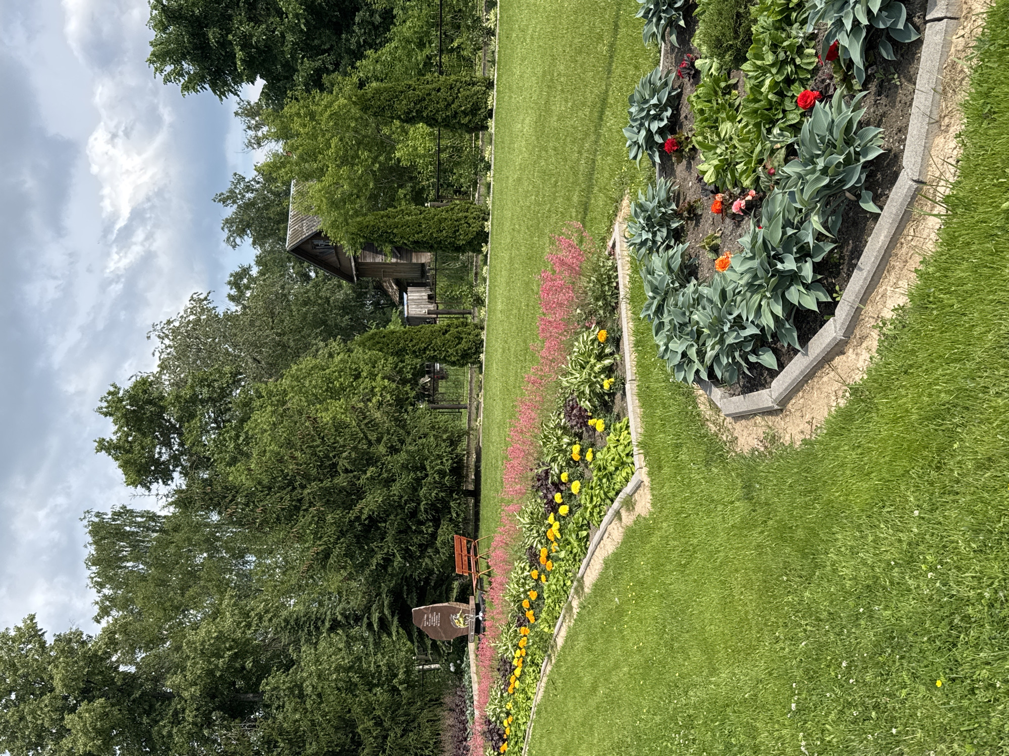This is a full, eye-level shot of a landscaped park area with a large flowerbed in the foreground, a lawn, and trees in the background. The flowerbed contains various plants, including hostas, marigolds, and other flowering plants in shades of pink, yellow, and red. The lawn is neatly mowed and appears to be well-maintained. In the background, there is a wooden building with a rustic appearance, a fence, and several tall evergreen trees. The sky is partly cloudy. This scene is located in the city of Chudovo, Russia, and appears to be a peaceful and well-kept public space, possibly a memorial park due to the presence of a large stone monument and a bench. There are no people in the image.