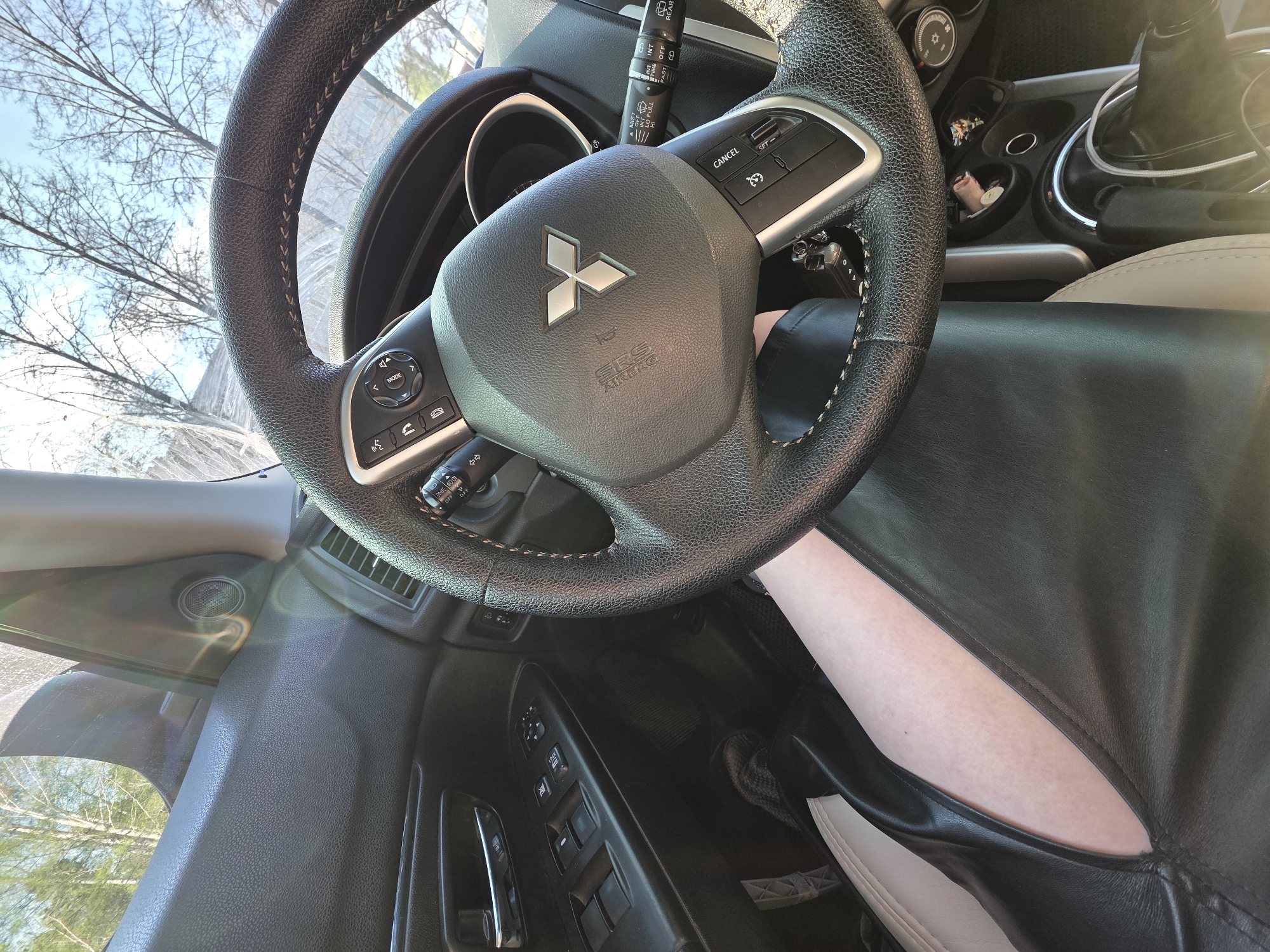 The image depicts the interior of a car, specifically the driver's seat area. The steering wheel, featuring a Mitsubishi logo, dominates the view. Controls and levers for functions like cruise control and windshield wipers are visible on the steering column. A person, presumably the driver, is seated in the car, with their leg visible against the dark leather-like upholstery. The car's door panel, with window and lock controls, is also in view. The background, visible through the windshield and side window, shows trees and a sky, suggesting an outdoor location. There is no information in the image that indicates it was taken in Chaikovsky, Russia.
