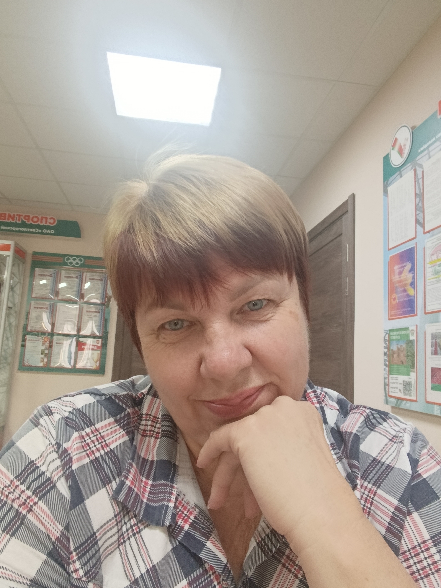 A middle-aged woman with short, blonde and brown hair and blue eyes is looking at the camera, resting her chin on her hand. She is wearing a red, white, and blue plaid shirt. The background appears to be an indoor office or public space, possibly a sports-related facility, as suggested by a display with Olympic rings and what looks like official documents or certificates. There is also a wall with multiple posters or information boards on it, one of which has a circular emblem that appears to be the flag of Belarus. The overall lighting is bright, with a visible overhead light fixture. The image appears to have been taken in Svietlahorsk, Belarus.