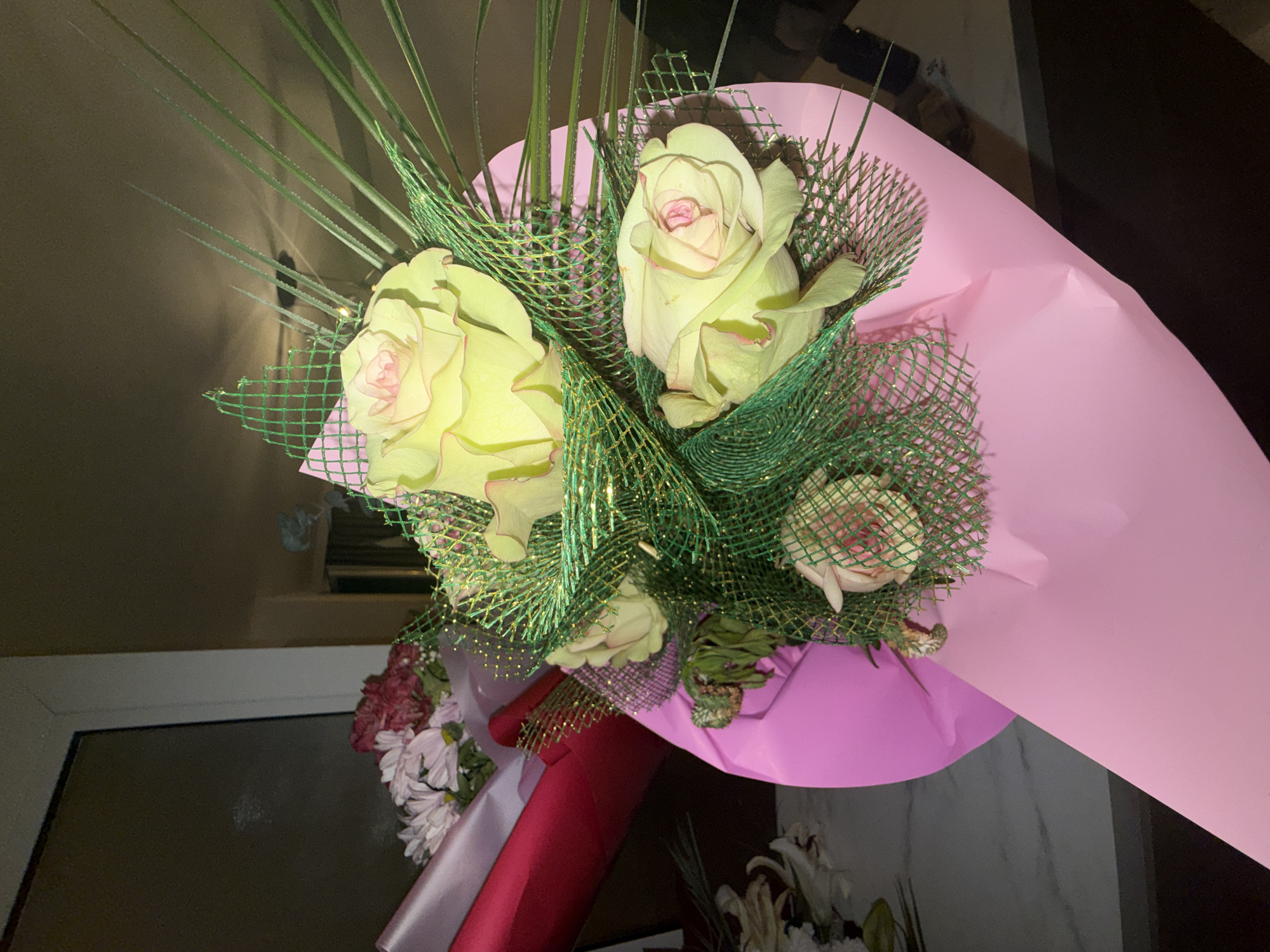The image shows a bouquet of flowers wrapped in pink paper and secured with red and pink ribbon. The flowers include yellow and pink roses with green accents and filler, along with other flowers in white, pink, and burgundy hues at the base. The bouquet is placed indoors, as suggested by the presence of a window frame and wall in the background. The specific location within Blagoevgrad, Bulgaria cannot be determined from the image. There is no evidence of people or interactions within the scene.