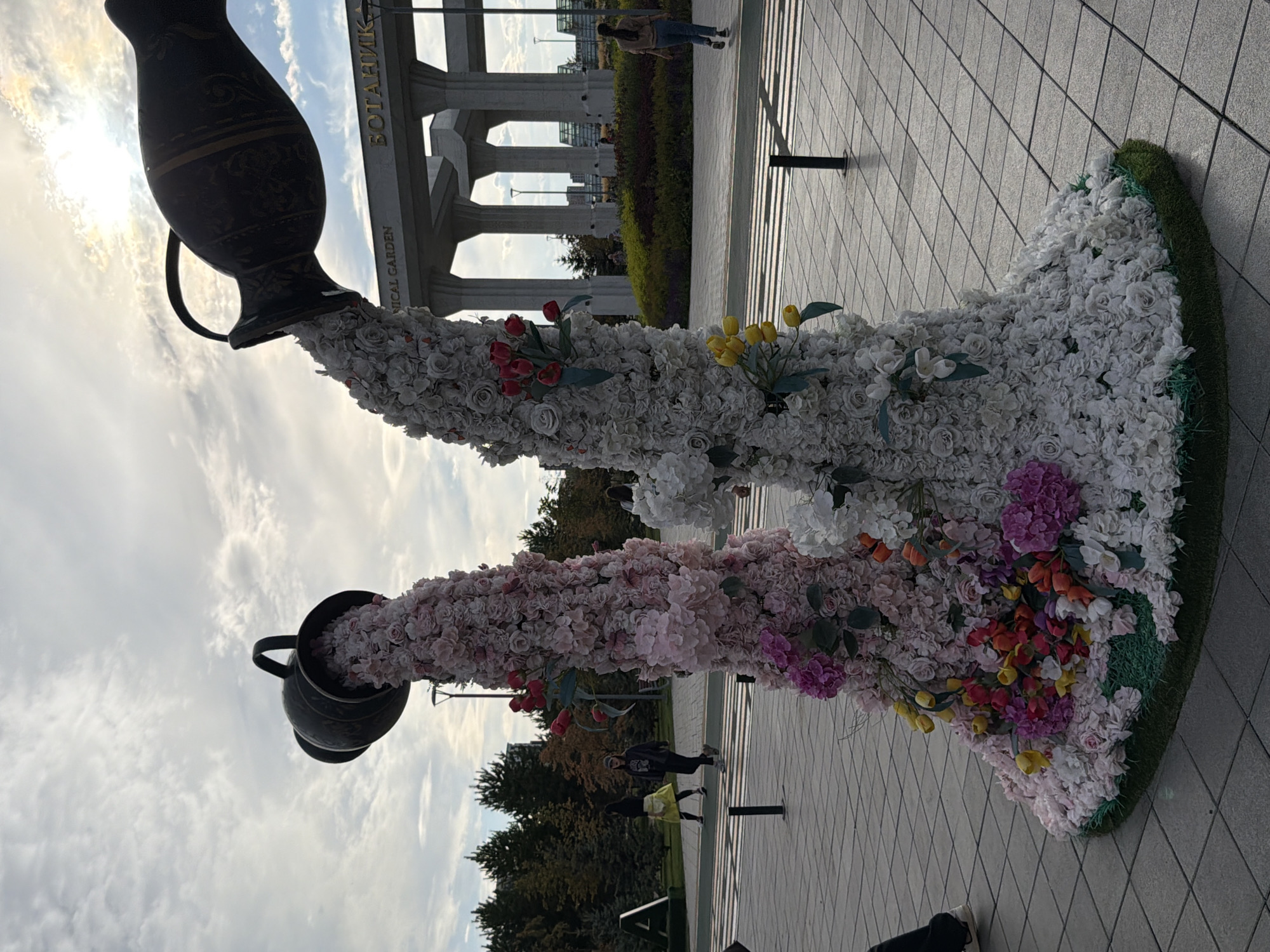 In Astana, Kazakhstan, an art installation featuring two large, dark-colored vases is prominently displayed outdoors. From each vase, a cascade of artificial flowers spills downwards, creating tall, cone-shaped structures. The left vase appears to be pouring pink and white flowers, while the right vase is showering white and light pink flowers. The base of each floral sculpture is adorned with additional flowers in various colors, including pink, yellow, and orange, and is surrounded by a green artificial grass base.
In the background, a modern building with large columns and an arched entrance is visible. A sign above the entrance reads "БОТАНИК" in Cyrillic, and below it, in English, "ICAL GARDEN," indicating the location is likely the Astana Botanical Garden. The sky is partly cloudy, with the sun visible through the clouds, casting a bright light.
Several people are present in the scene. In the middle ground, two individuals are walking across a pedestrian crossing. One person, on the left, is wearing a yellow skirt and dark top. The other person, to their right, is wearing a dark hoodie and pants. Further in the background, on the right side, a person in a brown top and blue jeans is walking away from the camera. The ground is paved with square tiles. The overall impression is a public outdoor space with an artistic floral display in front of an architectural landmark.