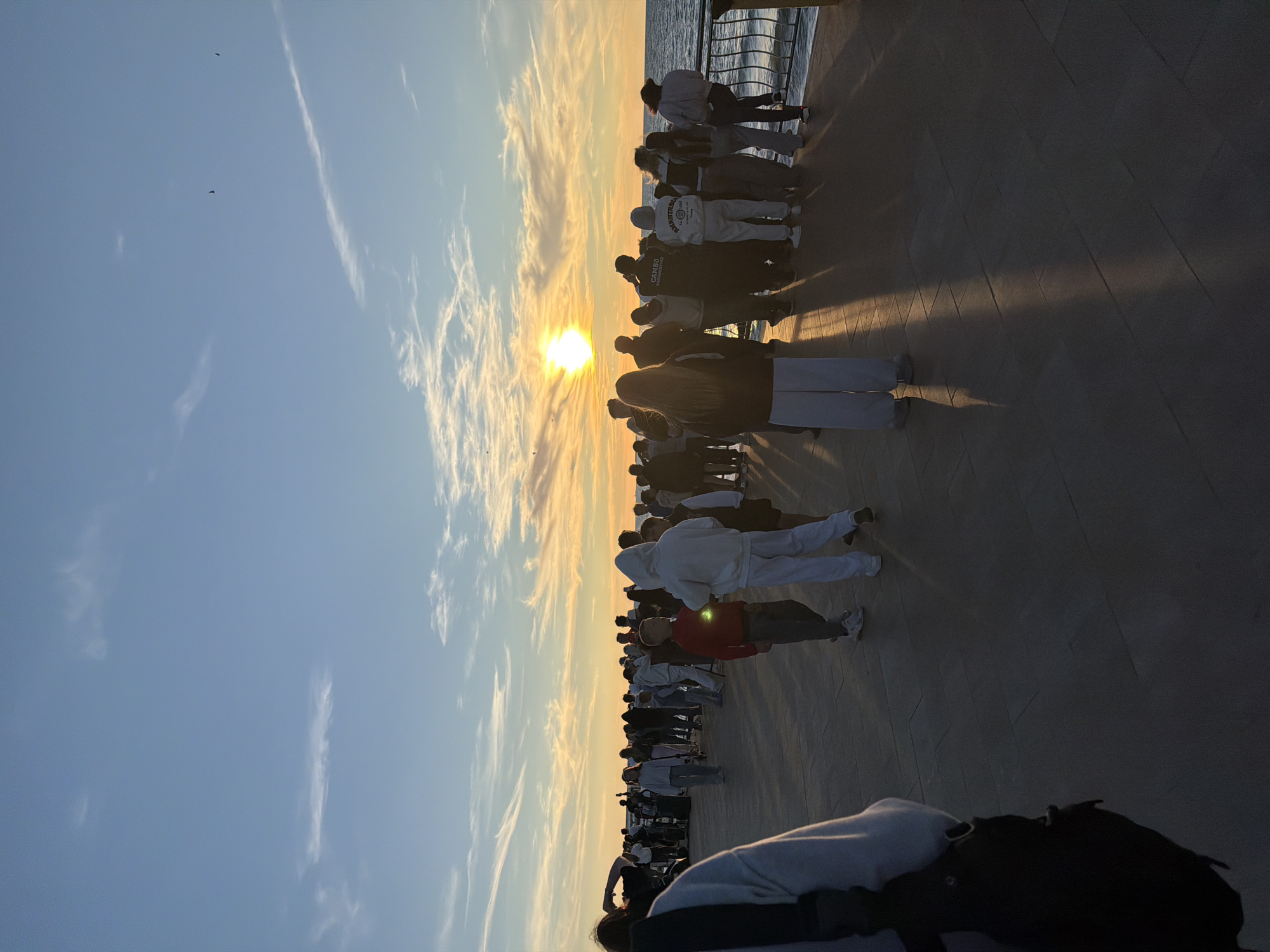 This is a vertical, full-shot photograph of a crowd of people gathered on a paved promenade by the sea during sunset. The sun is low on the horizon, casting long shadows across the promenade and illuminating the sky with warm hues of orange and yellow. Wispy clouds are scattered across a light blue sky. The sea is visible in the background on the right side of the image, with a railing separating the promenade from the water.
The crowd is diverse, with individuals of varying ages, many wearing casual clothing like hoodies and sweatpants. Some people are looking out at the sea, while others appear to be talking amongst themselves. In the foreground, a person wearing a light-colored hoodie and dark pants is partially visible from the back, with a black backpack on their left shoulder. To the right of the foreground individual, two young boys are present, one wearing a red shirt and dark pants, and the other in a white hoodie and light-colored pants. The promenade is paved with rectangular tiles, and the lighting suggests it's late afternoon or early evening. The scene is likely taking place on the promenade in Zelenogradsk, Russia, given the coastal setting and potential for local gatherings at sunset.