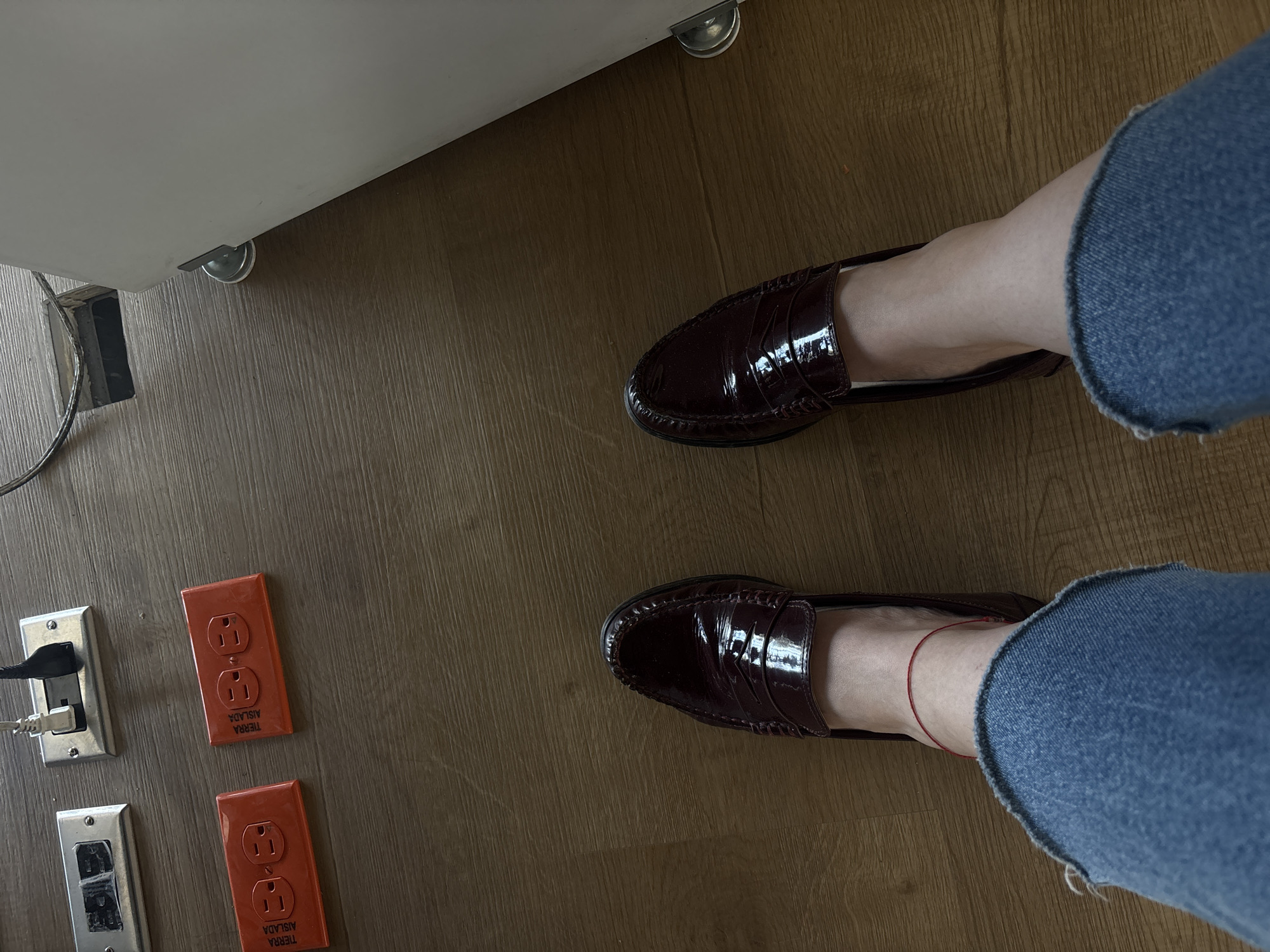 The image shows a downward-facing perspective of a person standing on a wood-look floor. The person is wearing blue jeans with frayed hems and dark burgundy loafers. Their ankles are visible, with a red string bracelet on one. To the left of the person's feet are three electrical outlets. Two of the outlets have orange faceplates. A white object with a silver castor wheel is partially visible in the upper left corner. A power cord extends from underneath this object. There is no information in the image to suggest the location of Buenavista, Mexico.