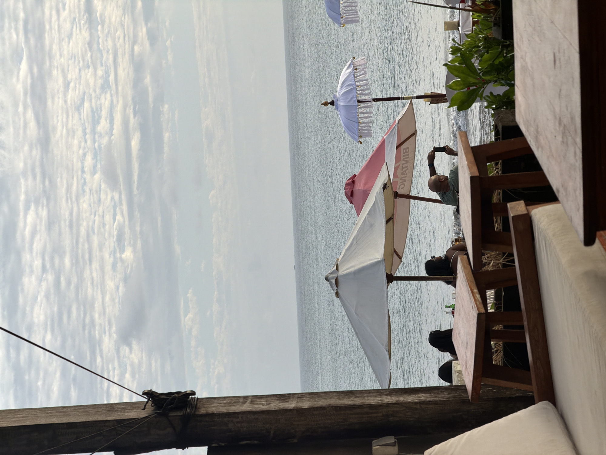 The image depicts a beachside scene at a cafe in Kuta, Indonesia, with a focus on outdoor seating and the ocean. In the foreground, two wooden tables are visible, one with a cushioned bench behind it. Further back, several beach umbrellas are set up. One is white, another is red and white with "BINTANG" visible on its underside, and a third is white with decorative fringes.
Several people are seated at tables under the umbrellas. Two women with dark hair are seated at a table. A man, bald with glasses, is sitting under the red and white umbrella, holding up a smartphone to take a photo. The ocean stretches out to the horizon under a cloudy sky. A small boat can be seen in the distance. Lush green foliage is present to the right of the scene, suggesting a tropical environment. The overall atmosphere is relaxed and indicative of a day at the beach.