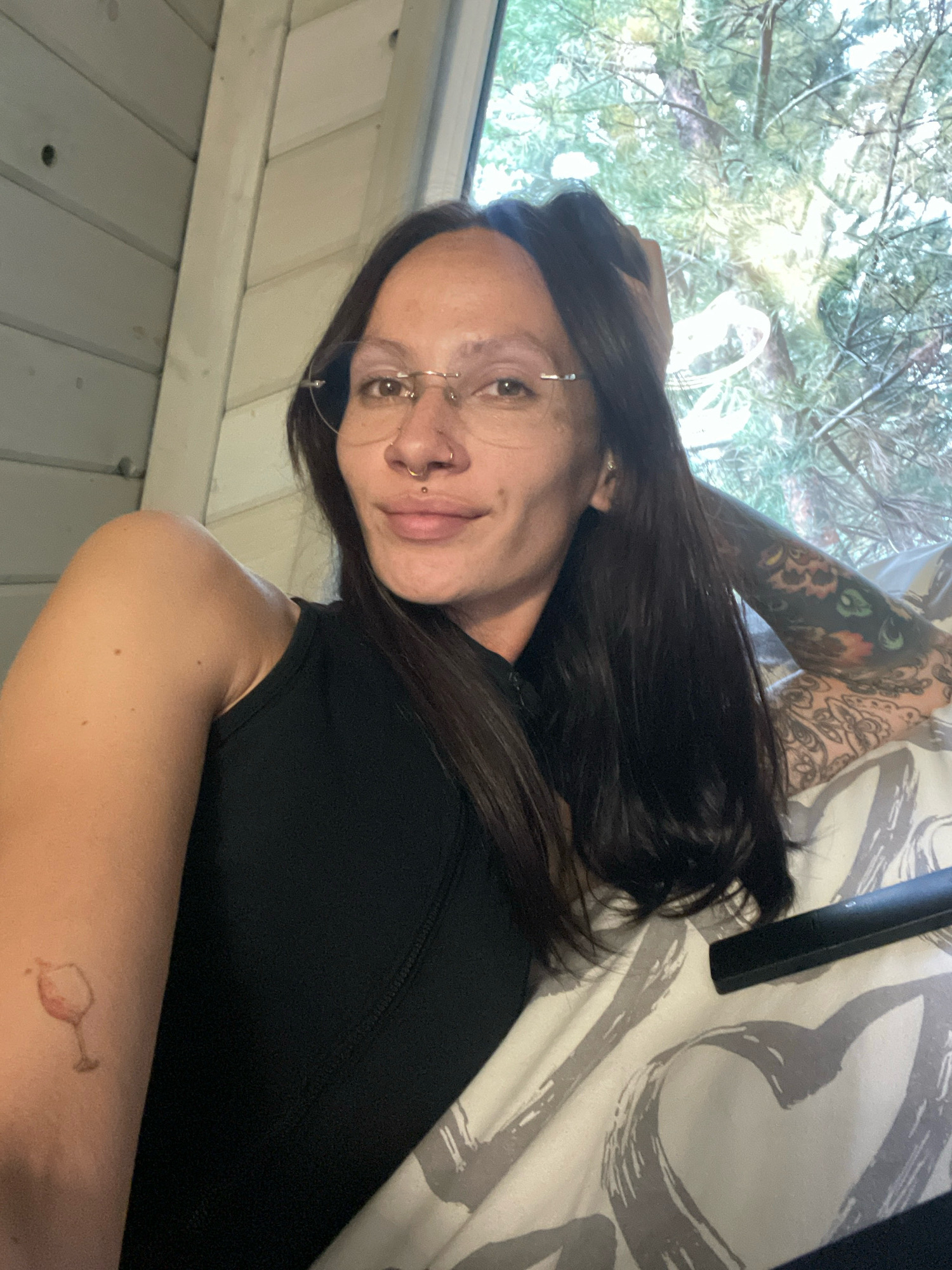 A young woman is lying on a bed and looking at the camera. She has long, dark hair, glasses, a nose ring, and a lip piercing. Her right arm is resting on her head, and her left arm is bent at the elbow, with a tattoo of a wine glass visible on her forearm. She is wearing a black tank top. The bed has a patterned duvet. Behind her, a window looks out onto trees. The room has wooden paneling on the wall to the left. The scene appears to be an indoor residential setting.