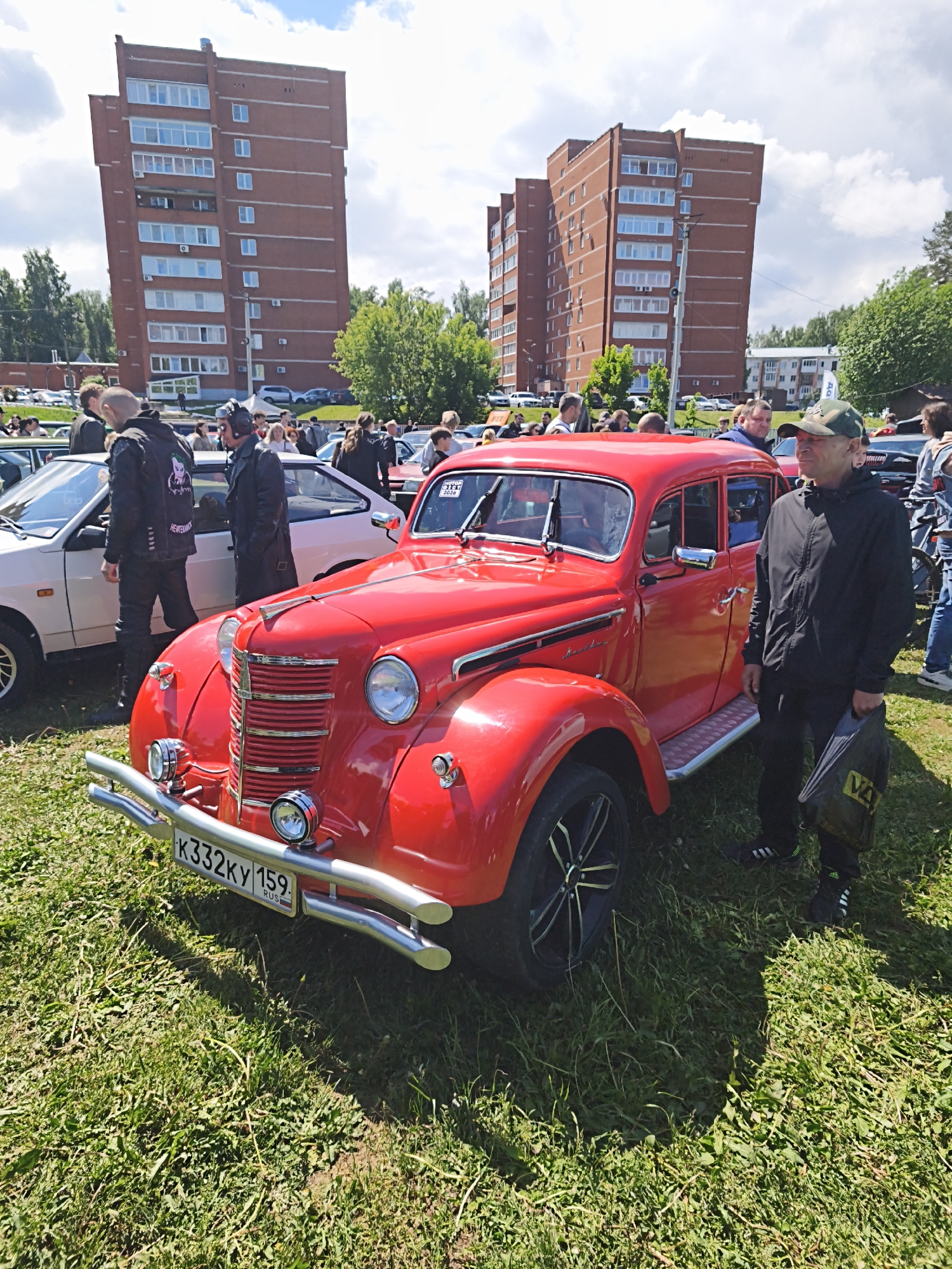 The image depicts an outdoor scene with a bright red vintage car as the main subject, located in Chaikovsky, Russia, as evidenced by the license plate. The car is surrounded by green grass, and in the background are two tall brick apartment buildings. A group of people are gathered around the car, suggesting a car show or public event. One man is standing next to the car, dressed in a black jacket, pants, and a camouflage cap. He's holding a black bag with yellow lettering. Other cars and people are visible in the background, along with some trees and a cloudy sky.