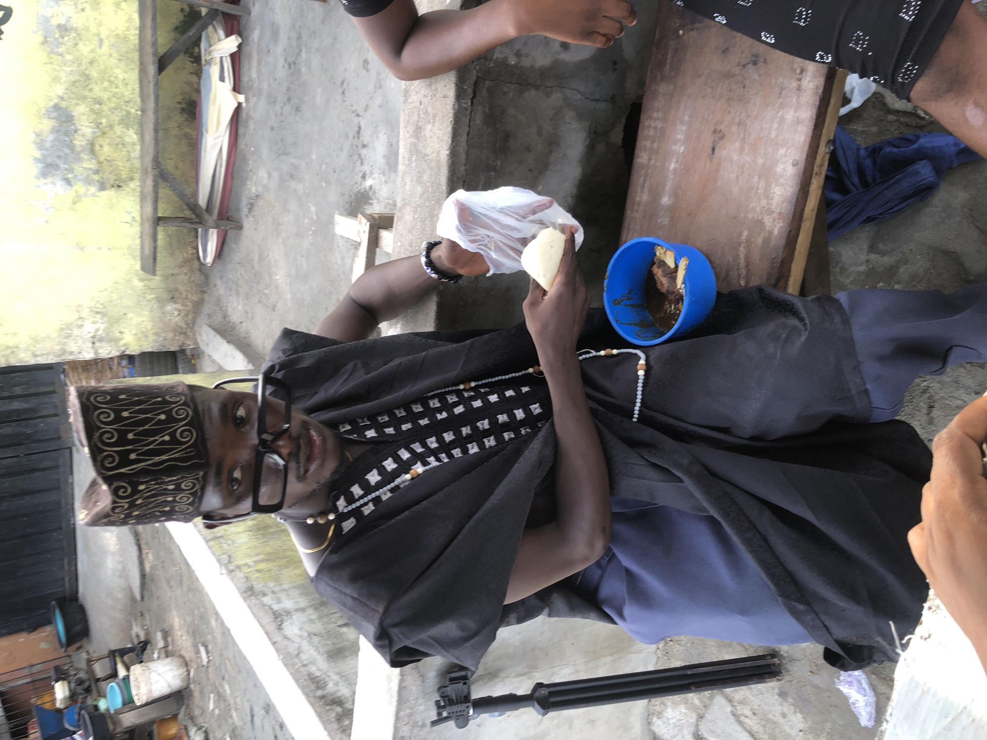 Ado, Nigeria: An individual in traditional Yoruba attire, including a patterned hat and flowing black robe, sits outdoors eating. A blue bowl containing food is on a small wooden table beside them. They hold a white food item from a clear plastic bag. Another person's arm is visible to the right, and another person is seated to the upper right. The ground is concrete. A metal tripod rests near the seated individual.