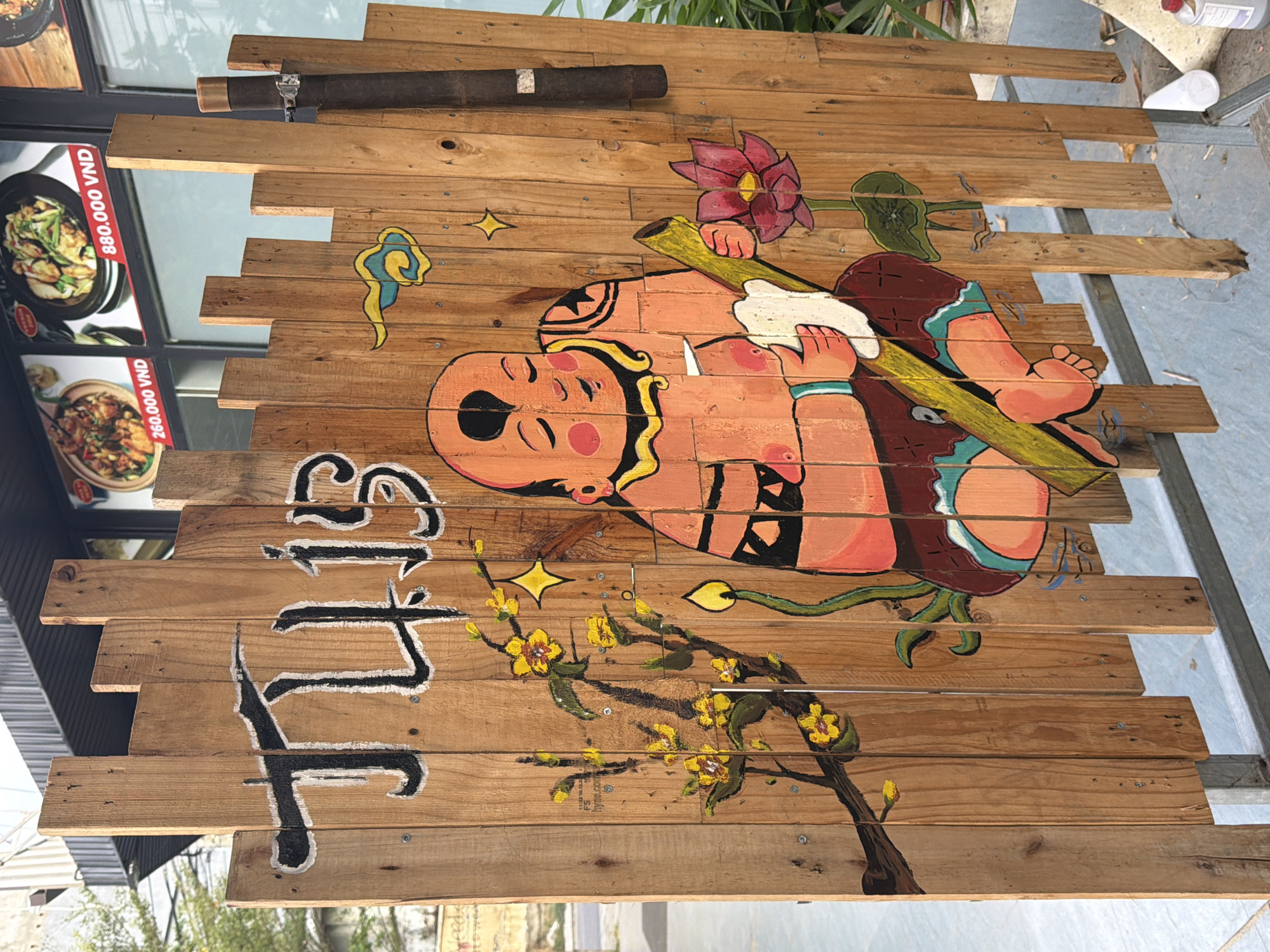 The image shows a wooden fence with a painting on it. The painting depicts a stylized bald figure in an orange tunic, holding a yellow stick and a piece of white cloth. The figure has a calm expression and is adorned with tattoos and jewelry. To the left of the figure is a branch with yellow flowers, and to the right is a pink lotus flower. Above the figure, there is a yellow and blue cloud motif, and a yellow star. The wooden fence itself is made of vertically oriented planks of varying heights, creating an uneven top edge. The fence is set against a backdrop that appears to be a shop or restaurant with windows displaying food and prices in Vietnamese currency (VND). Based on the context of the prices and the general aesthetic, this scene is likely located in the city of Đà Nẵng, Vietnam. The fence is supported by a metal frame at the bottom, suggesting it is a temporary or decorative structure. The environment appears to be outdoors, with some greenery visible in the background on the right side. The lighting suggests it is daytime.