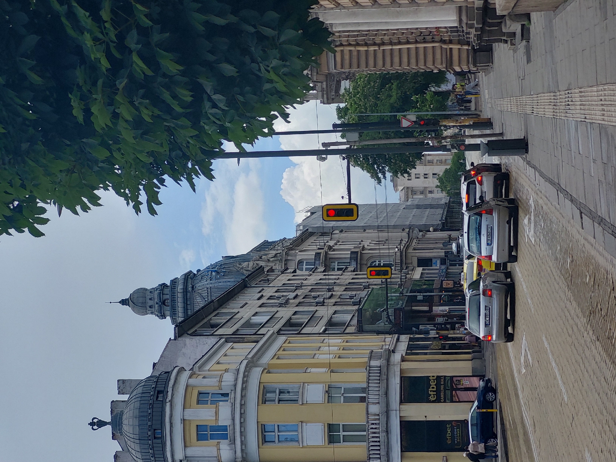 A street scene in Sofia, Bulgaria, shows buildings with domes and yellow facades. Traffic lights are visible, displaying red signals. A row of parked cars lines the right side of the street, with pedestrians standing near a dark-colored car. Green leaves from trees are in the upper left corner. The sky above is partly cloudy.