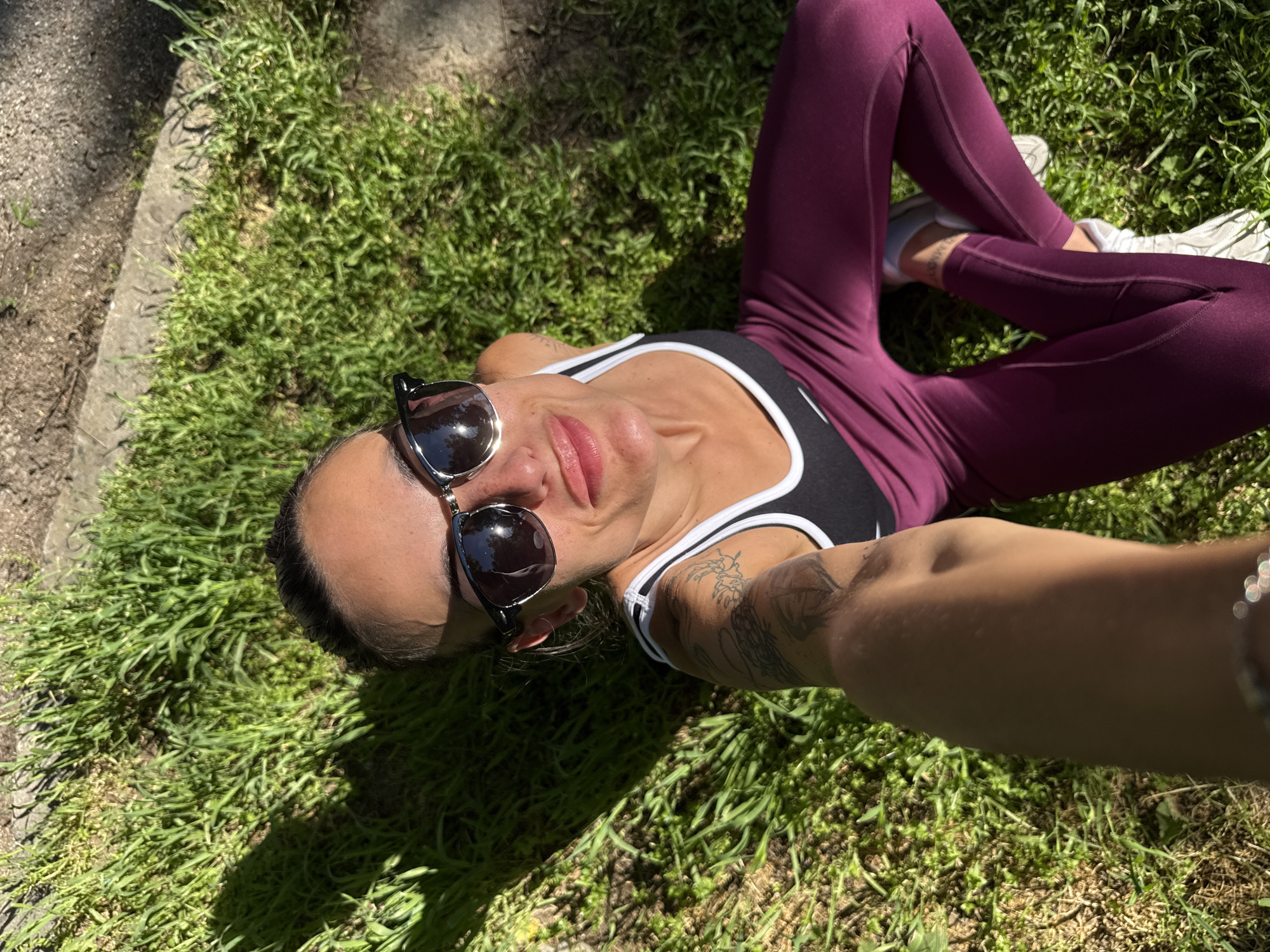 A woman lies on grass, wearing sunglasses, a black and white tank top, and maroon leggings with white sneakers. Her left arm is visible, showing a tattoo. The ground is covered in green grass and some dry leaves. There is a concrete edge visible in the background. The location is unknown.