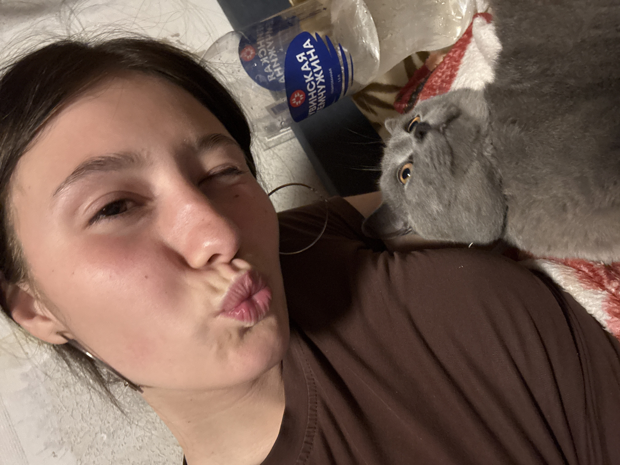 A young woman is lying down, puckering her lips into a kiss, with one eye closed, and looking directly at the camera. To her right, a gray cat with yellow eyes is resting on top of her brown shirt. Adjacent to the woman and the cat are a partially visible blanket and two empty plastic water bottles. The water bottle labels include Cyrillic text. The location is Фоки, Russia.