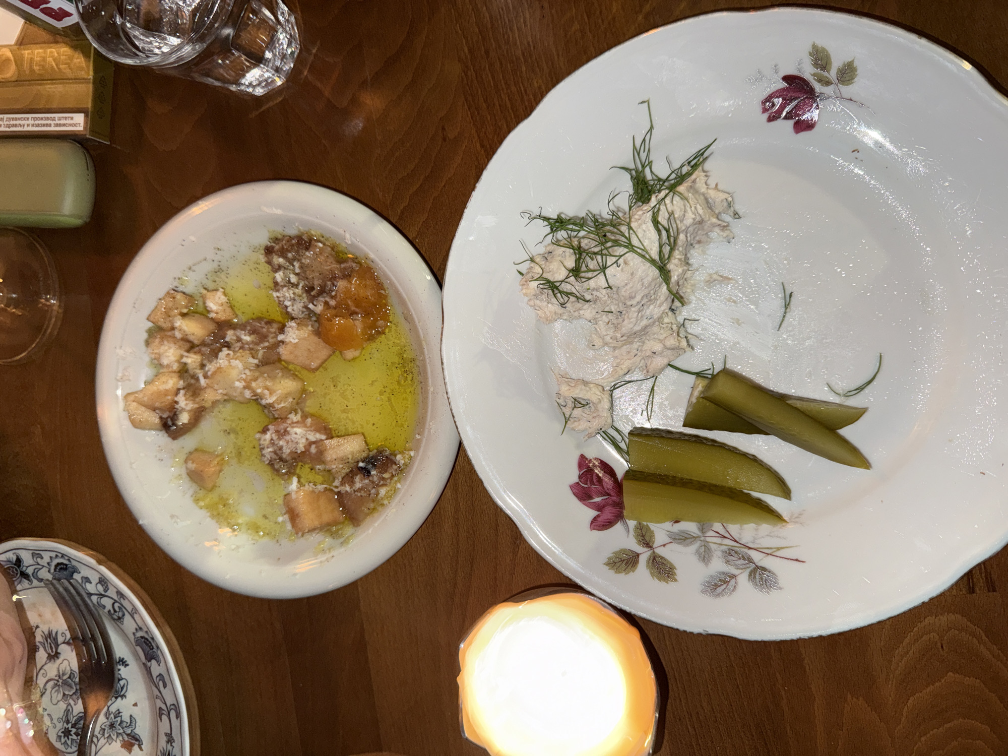 On a wooden table, two plates of food are visible, one containing a dish with what appears to be bread, cheese, and a yellow sauce, and the other with chopped pickles and a creamy spread garnished with dill. To the left of the plates, a lit candle in an orange holder emits a warm glow. In the background, a glass of water and what looks like a cigarette pack are present, suggesting a dining setting. The overall ambiance suggests a relaxed meal, possibly at a restaurant in Belgrade, Serbia.