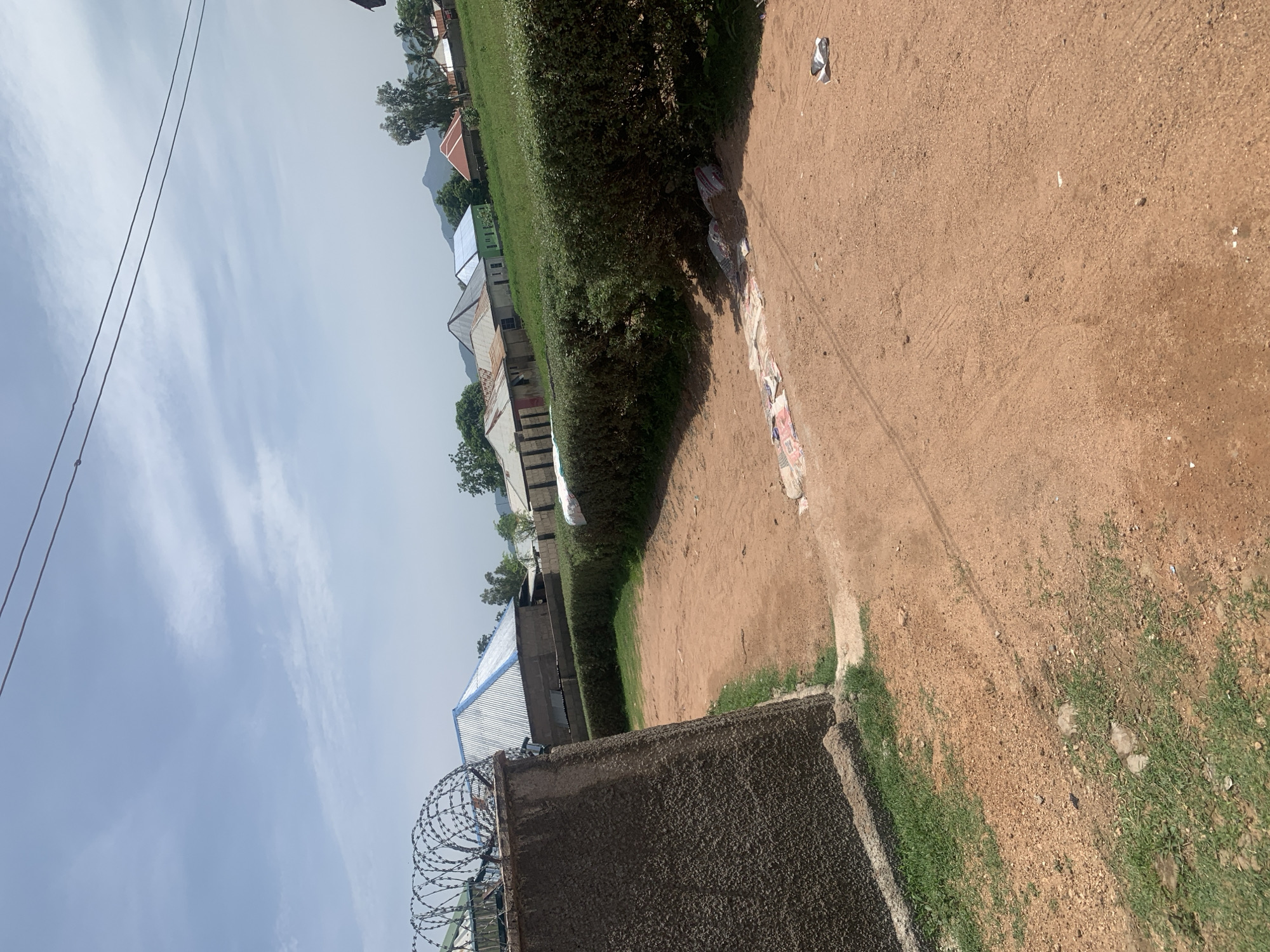 The image depicts an outdoor scene in Jauro Yinu, Nigeria, characterized by a dusty, reddish-brown dirt path or yard in the foreground. A rough, grey concrete wall, topped with barbed wire, runs along the left side of the frame. Adjacent to the wall is a patch of green grass.
Beyond the wall, a line of houses with corrugated metal roofs of various colors (silver, blue, red) are visible, situated on a grassy slope. Trees are scattered throughout the area, and in the background, distant hills or mountains can be seen under a partially cloudy sky.
The path itself shows some tire tracks. Scattered on the dirt path, particularly in the middle ground, are several pieces of debris, including what appear to be colorful paper or fabric items and some white plastic packaging. No people are present in the image. The overall environment suggests a rural or semi-urban setting.