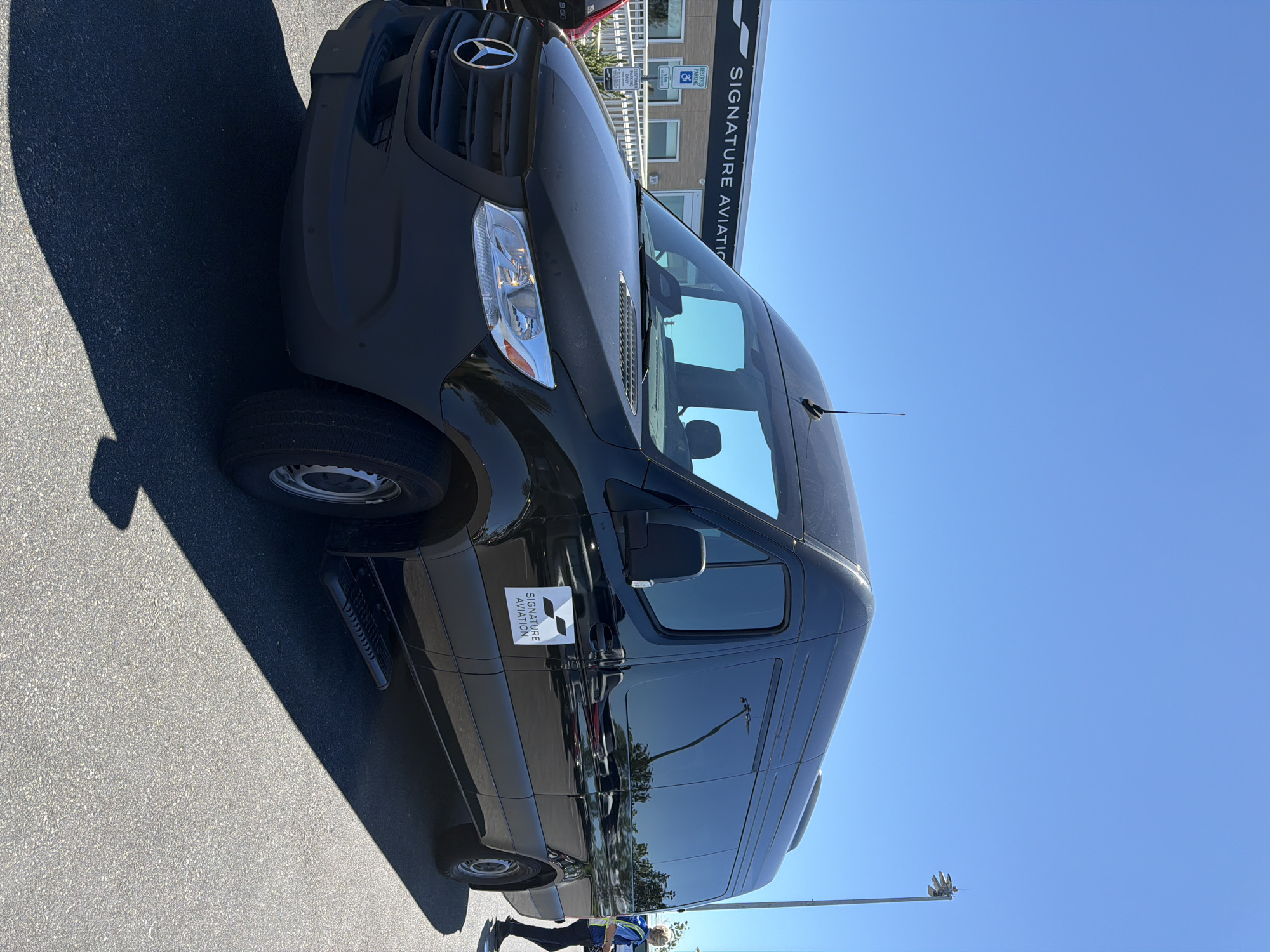 In Town of Southampton, United States of America, a black Mercedes-Benz Sprinter van is parked in an asphalt lot under a clear blue sky. The van has tinted windows and a "SIGNATURE AVIATION" logo on its side. In the background, a building with the same "SIGNATURE AVIATION" signage is visible, along with parking signs indicating reserved spots for disabled parking. A person in a safety vest and dark pants is walking away from the van towards the right side of the frame. The scene is illuminated by bright sunlight, casting a shadow from the van onto the pavement.