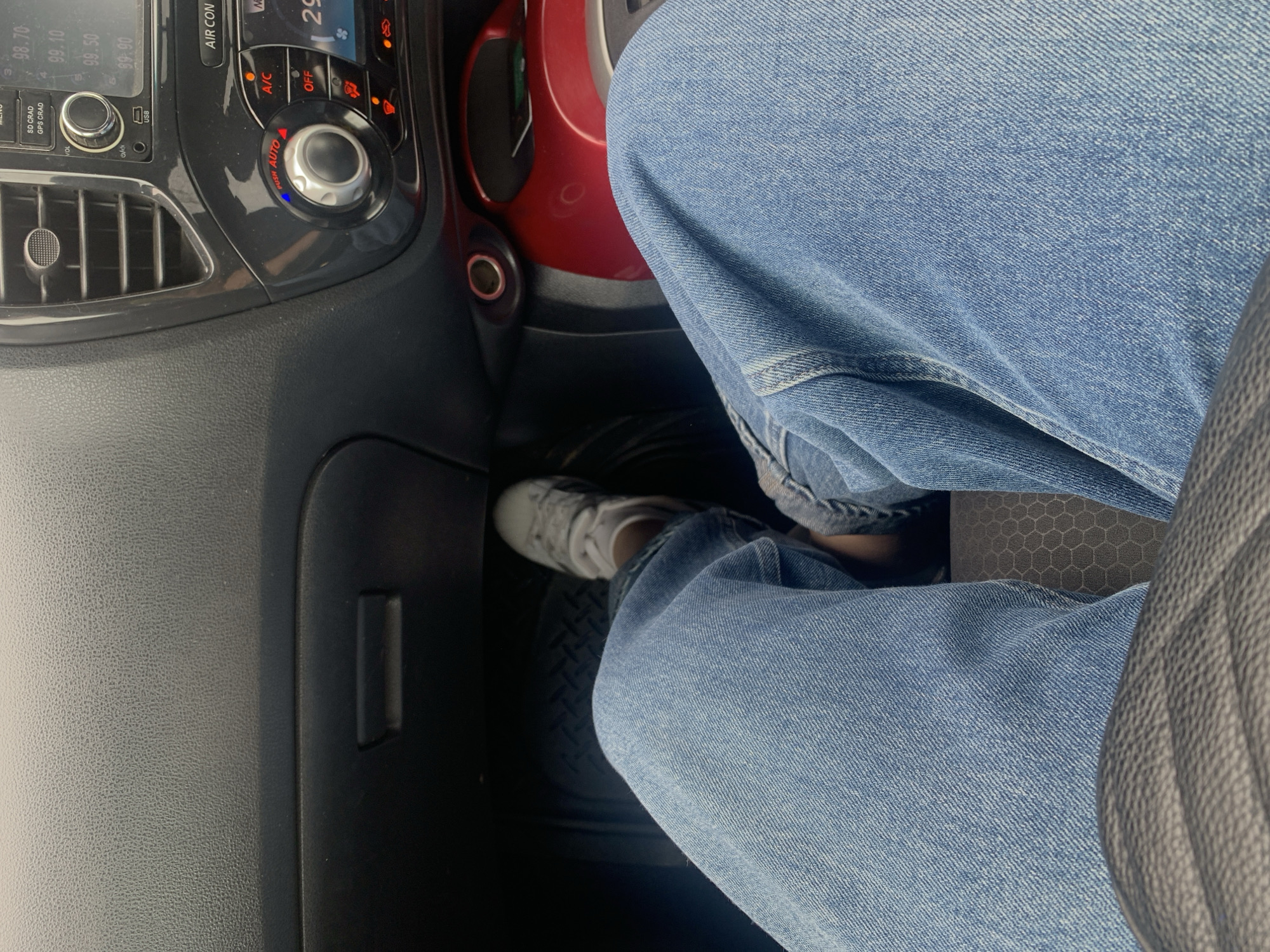 The image shows a close-up view of a person's legs and feet inside a car, focusing on the driver's side. The person is wearing light blue denim jeans and white athletic shoes. Their right foot appears to be resting on or near the car's pedals. The interior of the car is visible, including a portion of the dashboard with various controls, such as air conditioning settings and a radio display. The dashboard is dark gray and textured. A red-accented area is also visible to the right. The lighting suggests it is daytime. The image does not contain any information that would place it in Bratsk, Russia.
