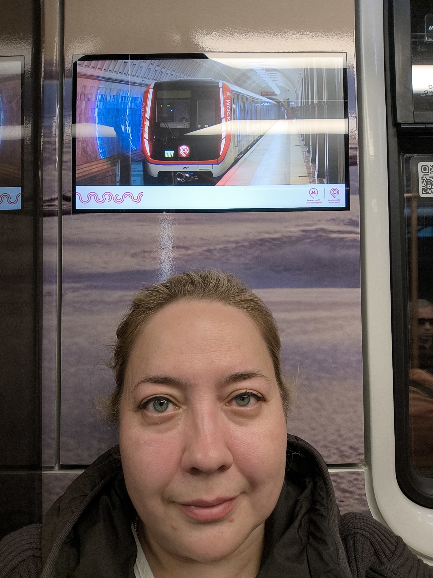 The image depicts a fair-skinned woman with light hair, wearing a dark-colored hooded jacket or sweater. She is looking directly at the camera. Behind her is what appears to be an informational display panel, with a video or image of a red and gray train in a subway station. The background of the panel shows a blurred, cloudy pattern in shades of brown and purple. Adjacent to the panel, on the right side of the image, is a narrow window or opening with a reflection of a person wearing sunglasses. The location is likely a subway station given the presence of the train image. The specific city and country are unknown.