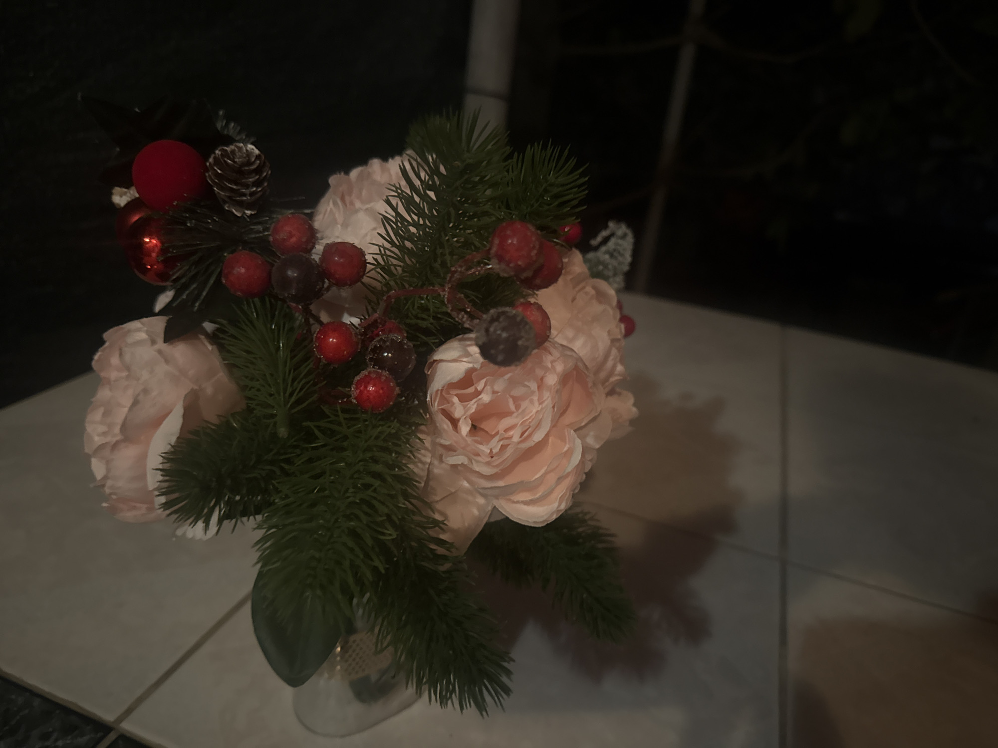 A close-up, slightly overhead shot shows a festive floral arrangement indoors. The bouquet features several large, pale pink peonies, interspersed with artificial pine branches and clusters of red berries. A small pine cone and a couple of shiny red ornamental balls are also visible among the foliage. The arrangement is presented in a metallic vase, partially obscured by the greenery. The scene is set against a dark background, with hints of a tiled surface beneath the vase. The lighting suggests it is evening or indoors, with soft illumination highlighting the textures and colors of the arrangement. The location is specified as Balaklavskyi District, Ukraine.