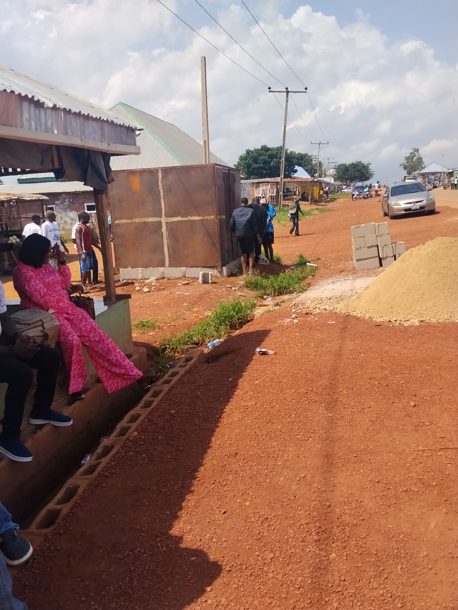 The image depicts a street scene in Durumi, Nigeria, characterized by red dirt roads and an overcast sky. In the foreground, a woman wearing a vibrant pink outfit is seated, looking to her right. Next to her, a person in dark clothing is seated, facing forward. In the middle ground, several individuals are standing and walking on the dirt road. To the left, a corrugated metal structure with an overhang creates shade. Further down the road, various stalls and buildings line the street, and a silver car is parked on the right. Construction materials, including a pile of sand and concrete blocks, are visible in the foreground and middle ground, suggesting ongoing development or repairs. The overall impression is that of a busy, dusty urban environment.