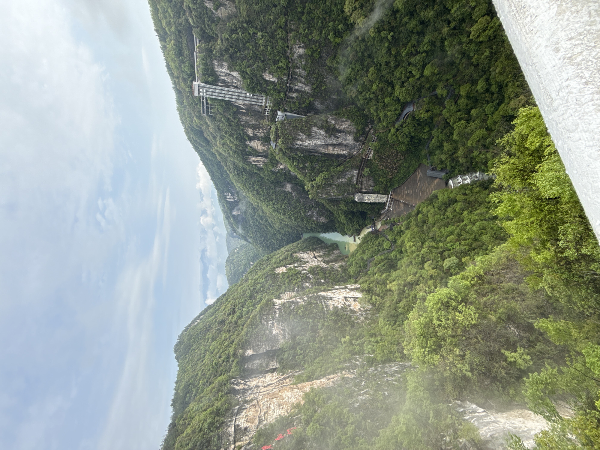 The image depicts a mountainous landscape in Zhangjiajie, China. Lush green vegetation covers the slopes. A river flows between the mountains. There are structures built into the mountainside, including a tall lift (elevator) and a concrete dam. The sky is partly cloudy. A waterfall cascades down the cliff in the foreground.