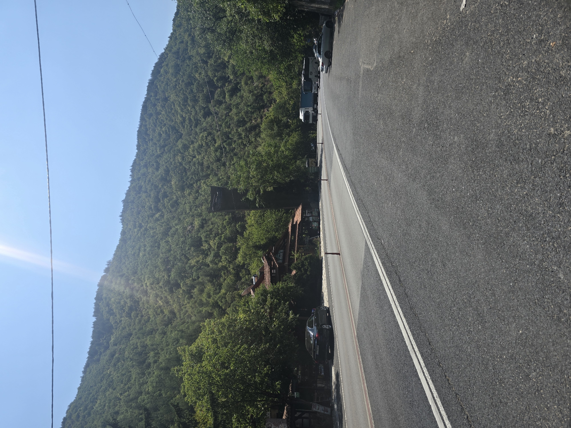 The image shows a road curving towards the right, with a steep, densely forested hill rising on the left side. Several vehicles are parked along the right side of the road, including a white tour bus, a silver sedan, and a white truck with a blue trailer. Further down the road, on the left side, there is a building with a red-tiled roof, possibly a restaurant or shop, with a dark sedan parked in front of it. The sky is clear and blue, with bright sunlight casting a glare. A power line runs horizontally across the top of the frame. The scene appears to be located near Stara Kresna, Bulgaria, given the mountainous terrain and architecture.