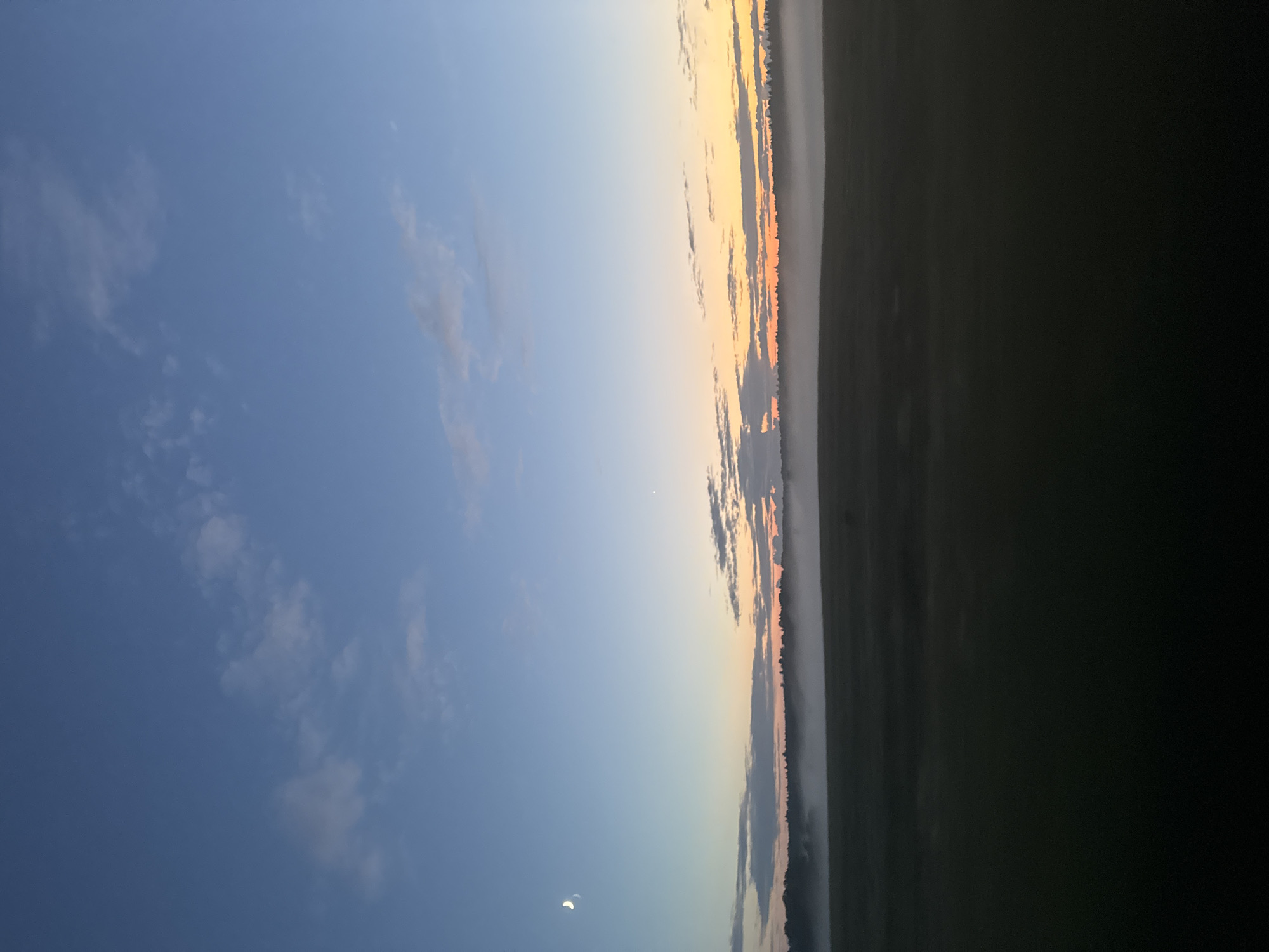 This is a wide shot of a field at dusk. The sky transitions from deep blue at the top to a pale orange near the horizon, with scattered clouds. A sliver of the moon is visible in the upper left corner. A thin layer of fog hovers over the dark, indistinct landscape of fields and trees that forms the lower half of the image. There are no people or animals visible in the scene. The location is identified as Егорье, Russia.