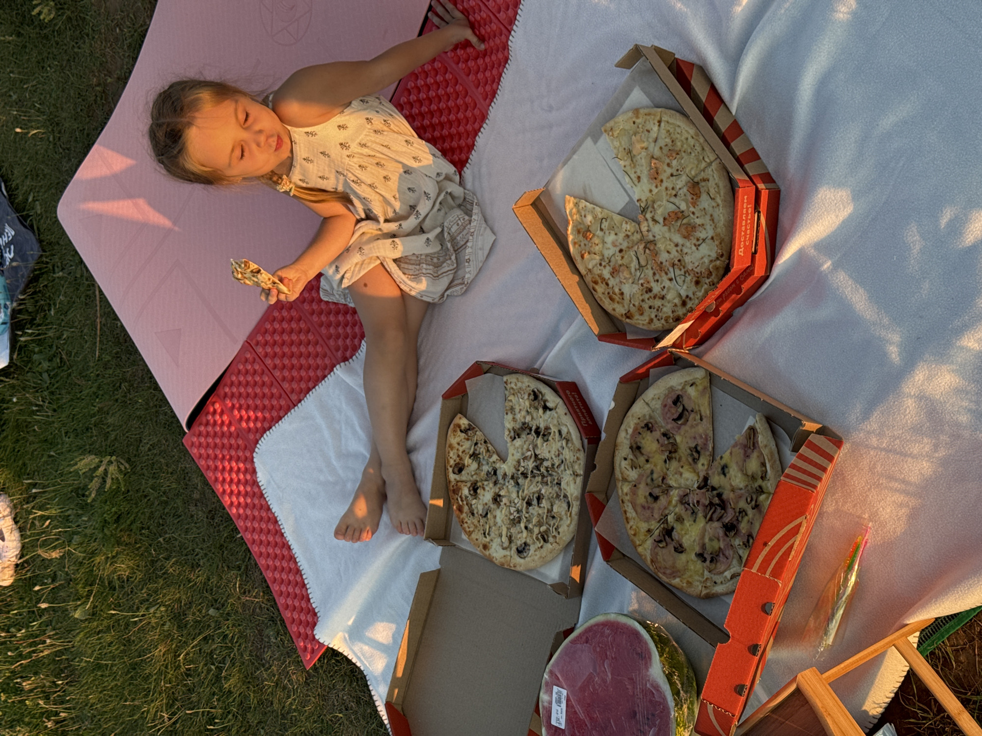 A young girl with blonde hair in pigtails sits on a white blanket spread out on the grass, eating a slice of pizza. She is wearing a light-colored dress. In front of her are three pizza boxes, two of which are open and contain pizzas. A half of a watermelon, wrapped in plastic, is also on the blanket. A red and pink mat is placed on the grass behind the girl. The scene appears to be a picnic. The location is the city of Olkhovka, Russia.