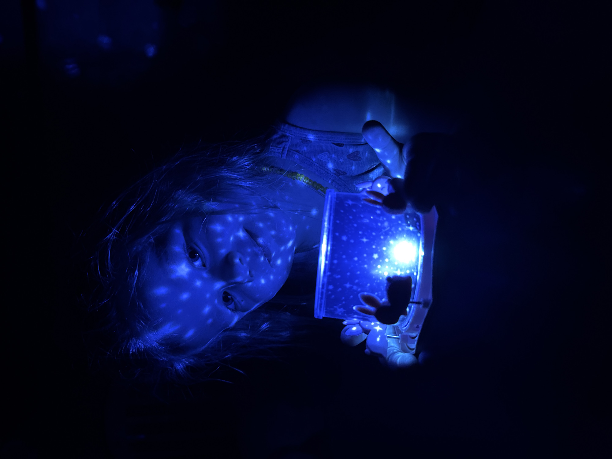 A young child is depicted in a dark indoor environment in Gornyi (Dvurechenskoe S/P), Russia. The child, appearing to be female with light-colored hair, is positioned centrally, gazing forward towards the viewer. She is illuminated primarily by an intense blue light originating from a cylindrical or cube-shaped light projector held in both hands. The blue light casts distinct white star-like patterns across her face, hair, and upper body. She wears a light-colored tank top with faint, dark patterns (possibly hearts) and a thin necklace or cord around her neck. The projector's upper surface features small figurines, including a prominent dark silhouette resembling a rabbit and another lighter-colored, less defined figure, possibly an animal. The surrounding environment is almost entirely dark, with the blue light and projected patterns forming the sole illumination.