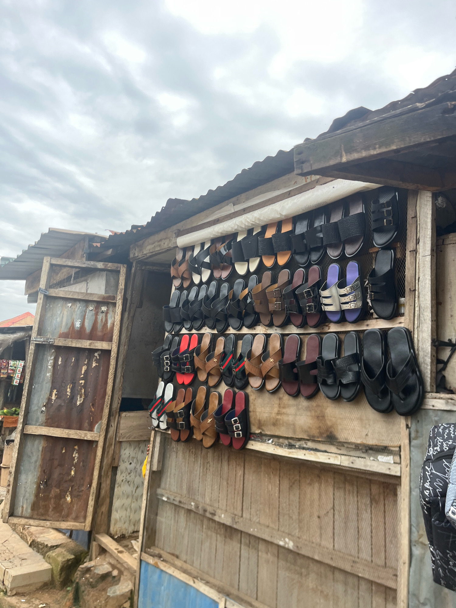 This image depicts an outdoor stall in Jos, Nigeria, displaying a variety of sandals and flip-flops. The stall has a corrugated metal roof supported by weathered wooden beams. The merchandise is arranged on shelves made of wooden planks. The sandals are predominantly black, brown, and some have red or patterned straps. Some sandals are made of leather, while others appear to be synthetic. To the left of the stall, a rustic wooden door with a metal mesh insert is partially open, revealing more of the stall's interior. A stack of what appear to be fruits is visible at the far left of the image. The ground in front of the stall is rough and appears to be a dirt or gravel surface. The sky is overcast with clouds. No people are clearly visible in the image, but the context suggests a market setting.